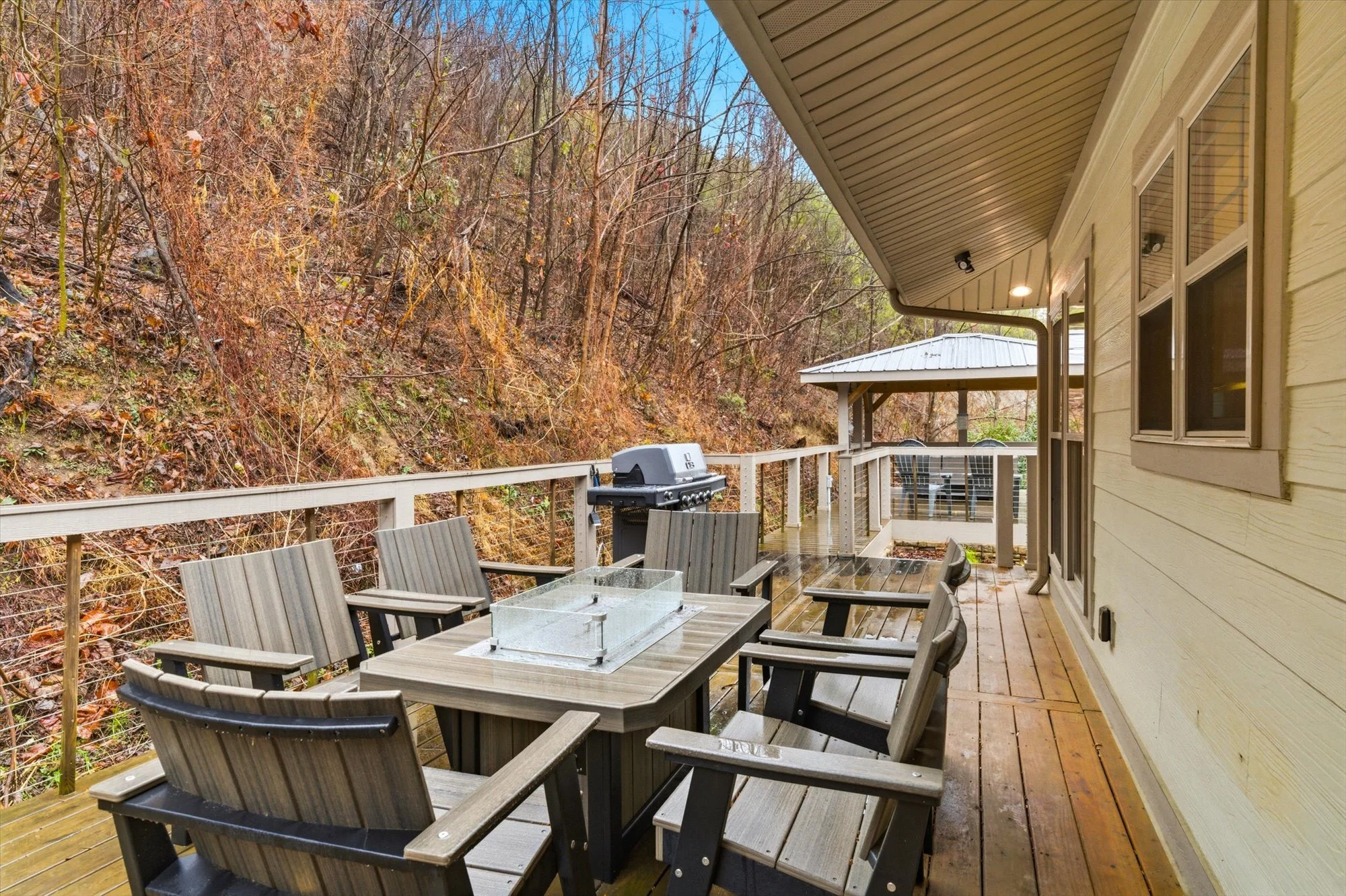 Outdoor wooden deck with a table and six chairs, a gas grill, and a covered gazebo in the background, overlooking a wooded hillside.