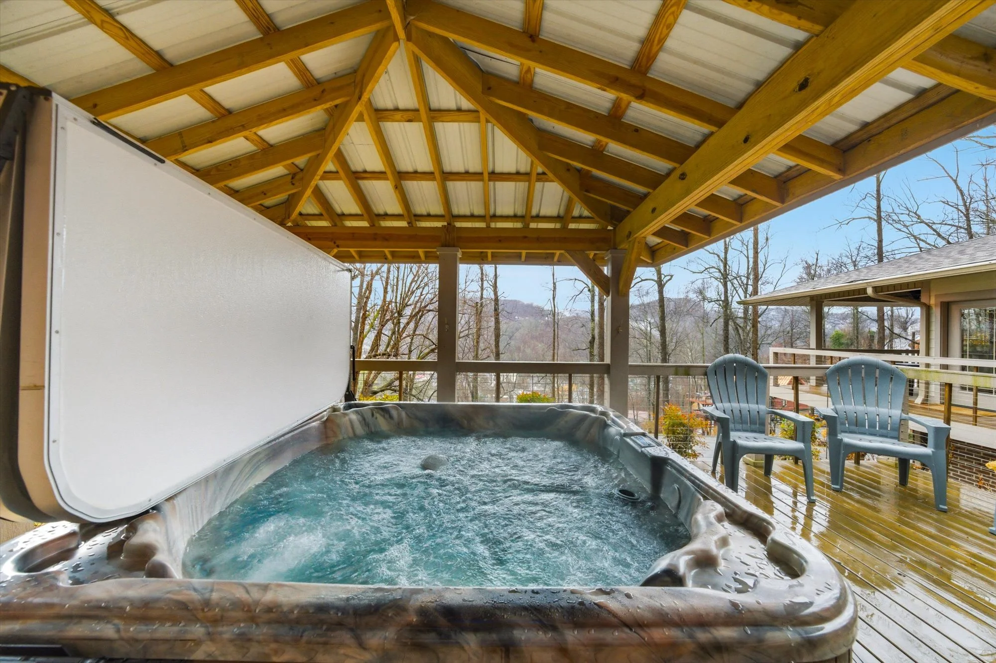 Outdoor deck with hot tub and two chairs, covered by a wooden roof, overlooking a wooded area.