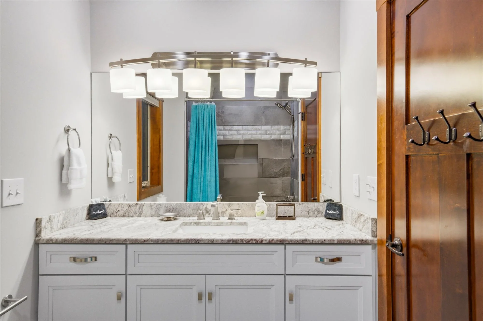 Bathroom vanity with white cabinets, granite countertop, and large mirror. There are two towel rings with white towels, a soap dispenser, and small makeup containers. The room has wooden accents and a view of a shower with a teal curtain and gray til