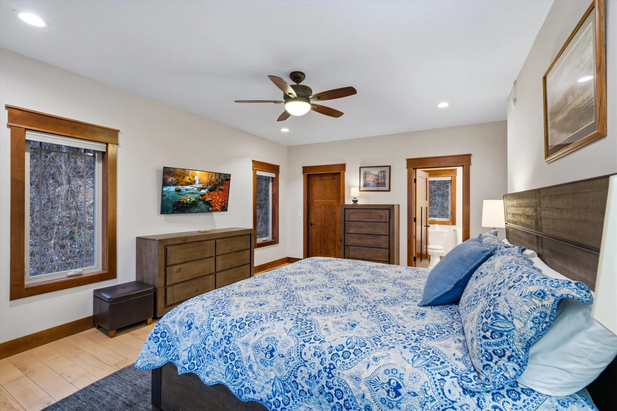 A bedroom with two windows with wooden trim, a wooden dresser, a wall-mounted TV displaying nature, a ceiling fan with blades, a bed with a blue and white patterned quilt, and a small ottoman. An open door leads to a bathroom.