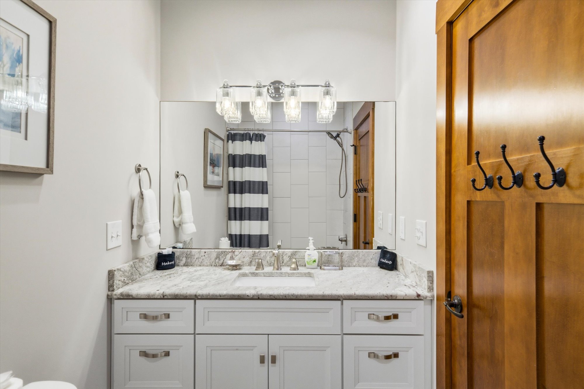 Bathroom vanity with white cabinets and granite countertop, large mirror, framed artwork, towel rings with white towels, soap dispenser, and toiletries; shower with striped curtain visible in the reflection; wooden door with hooks.