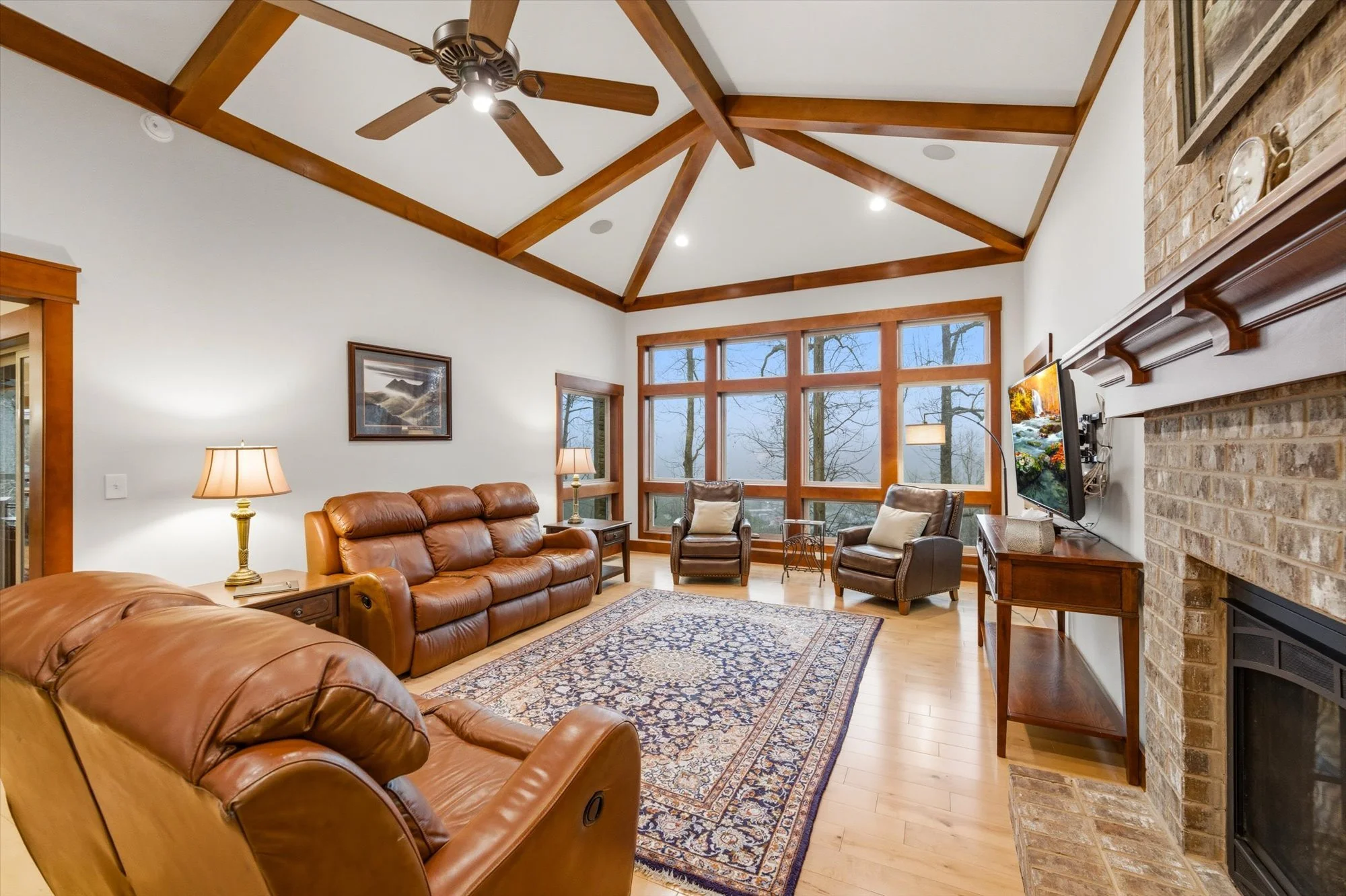 Living room with leather sofas, wooden chairs, a large window, brick fireplace, and ceiling fan.