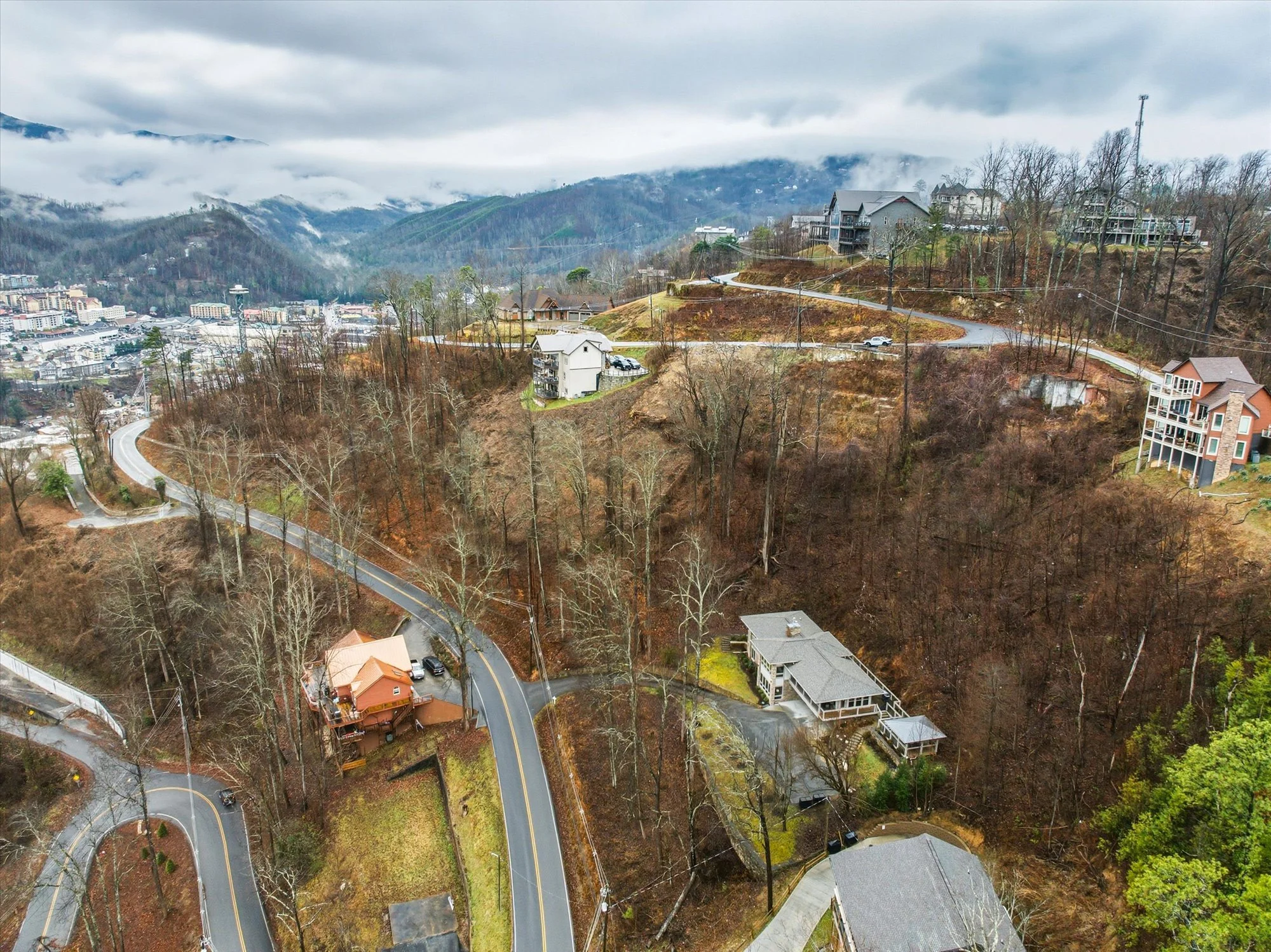 Hilly residential area with winding roads, houses, and trees, with mountains and clouds in the background.