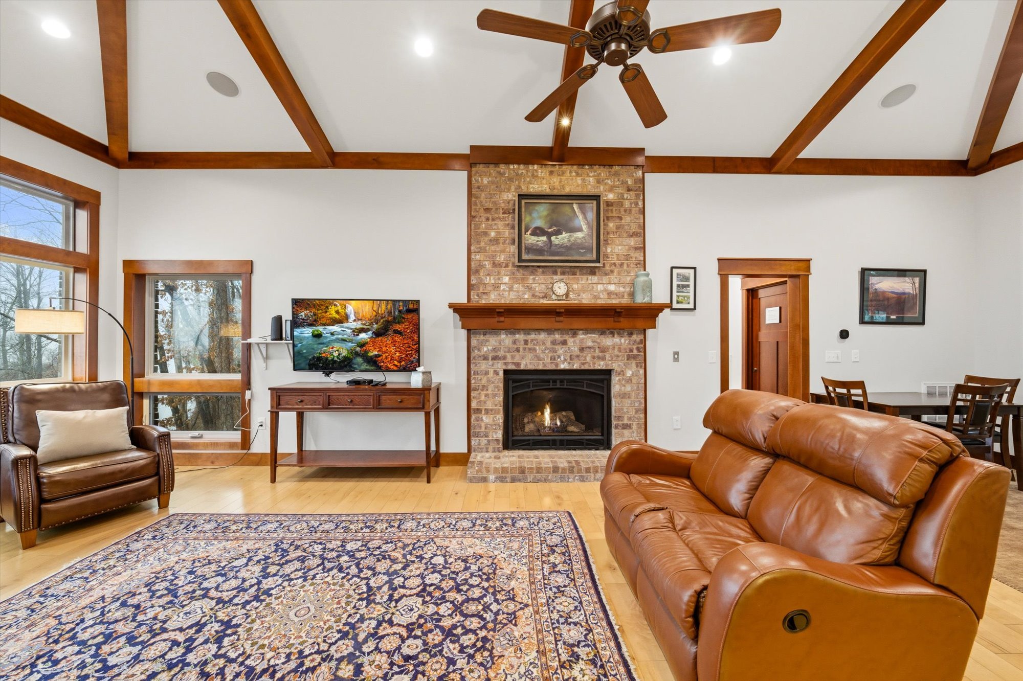 Living room with a brick fireplace, a leather sofa, a brown armchair, a wooden coffee table, a flat-screen TV, a patterned area rug, windows, and a wooden ceiling fan.