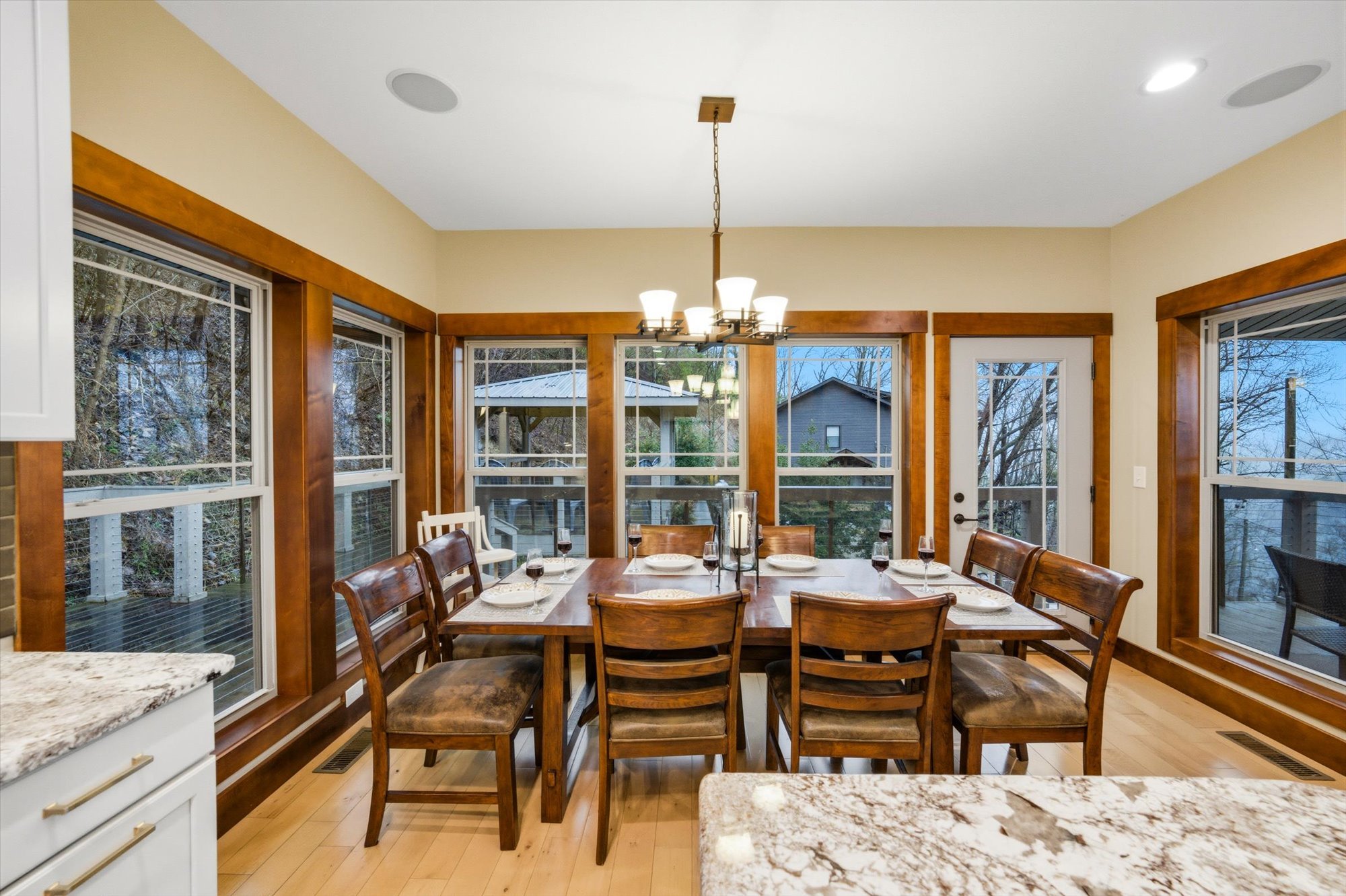Dining room with a wooden table, six chairs, large windows, and a door, decorated for a meal with plates, glasses, and a centerpiece, overlooking an outdoor deck and trees.