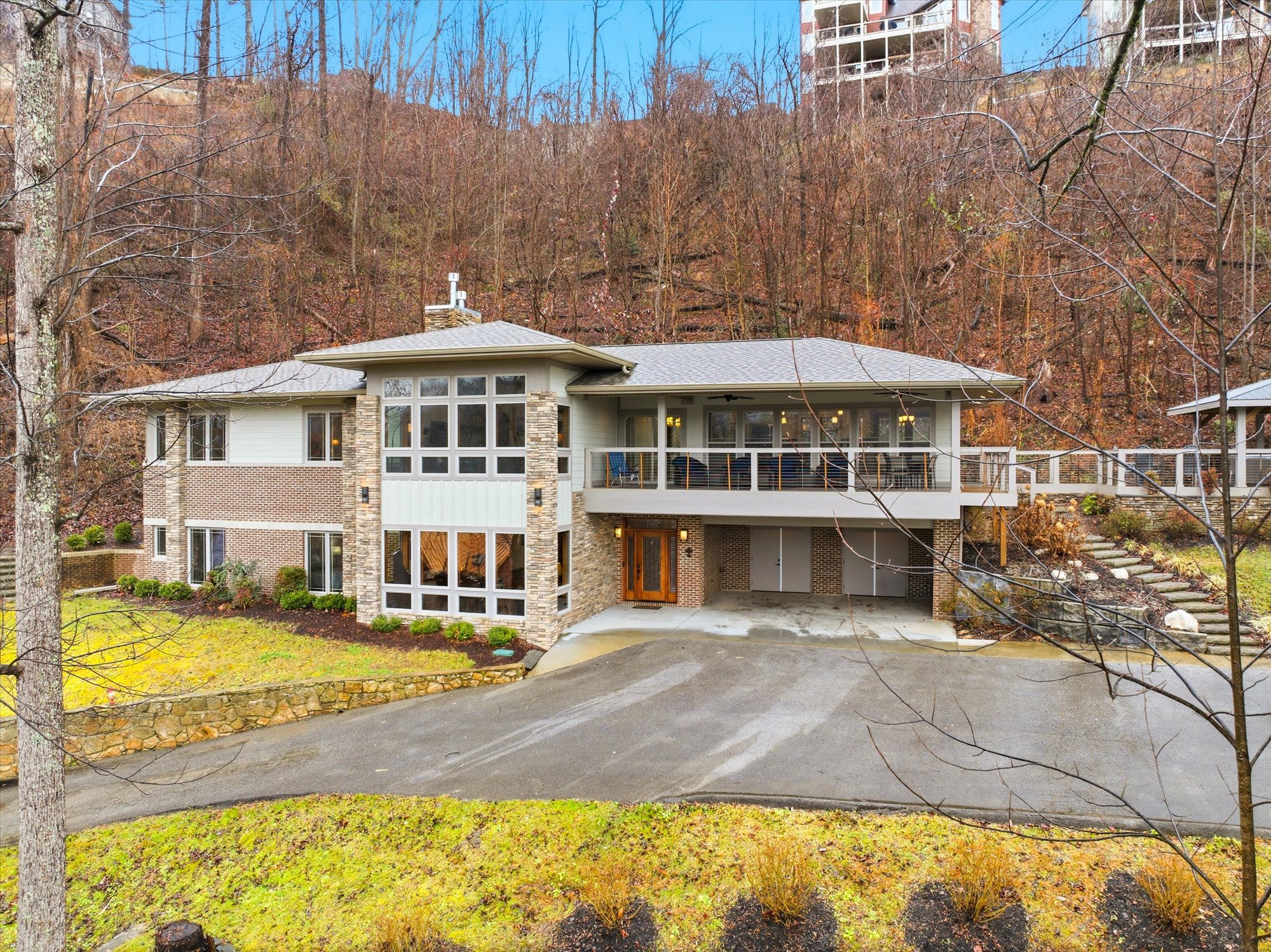 Modern two-story house with brick and siding exterior, large front windows, and a spacious balcony. The house is surrounded by landscaped yard with moss-covered ground, shrubs, and a driveway leading to a garage, set against a wooded hillside with le