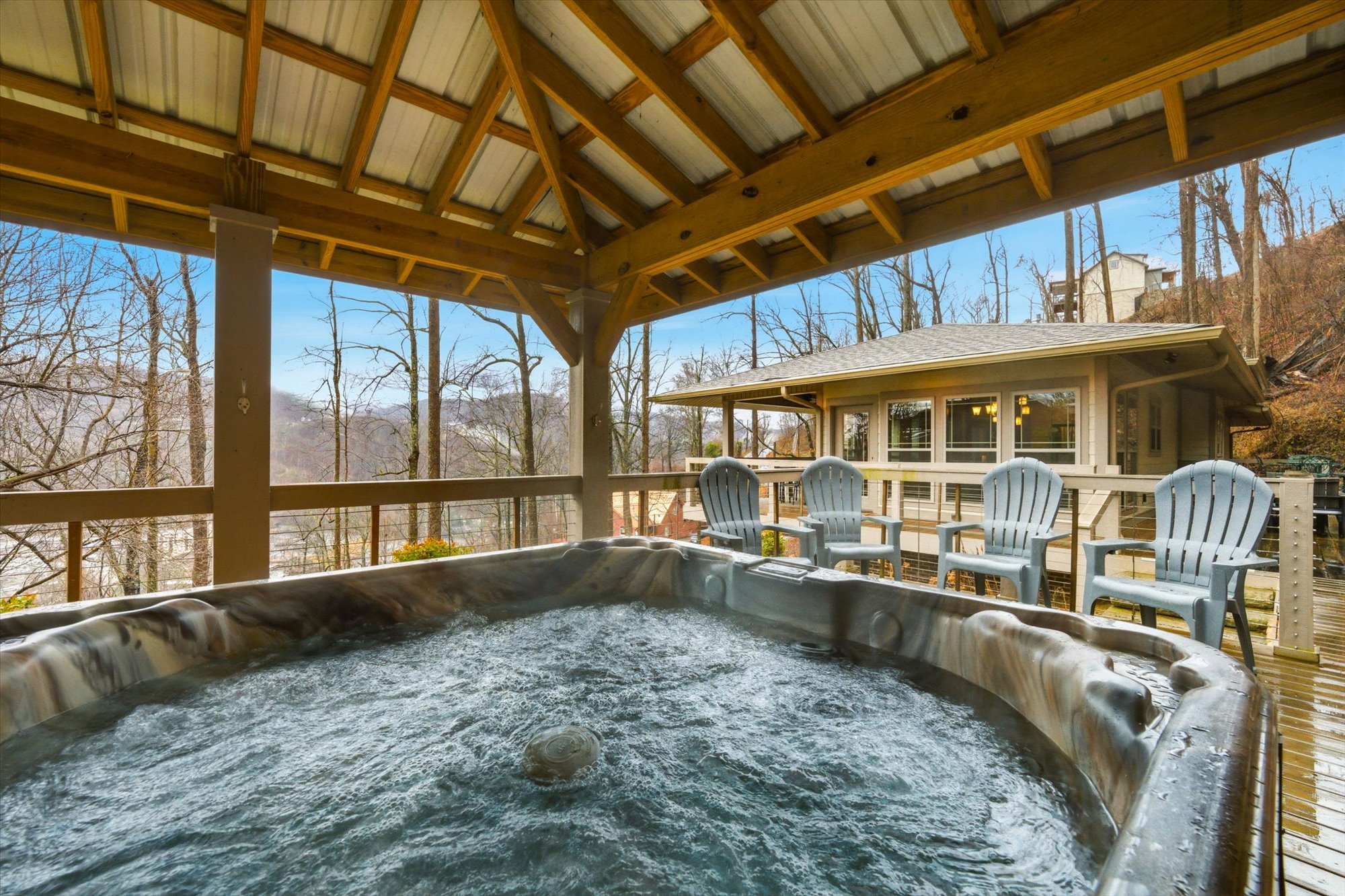 Outdoor covered deck with a hot tub, four blue chairs, and a view of a house through leafless trees.