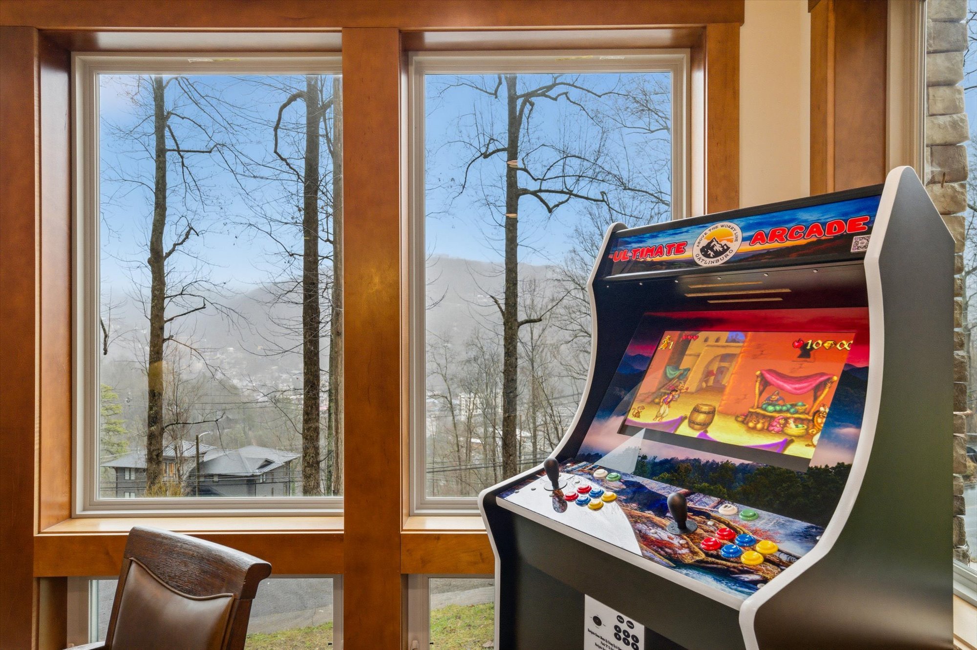 An arcade game machine with colorful controls and a screen showing a game scene, positioned next to large windows revealing a tree outside and a house in a wooded landscape.