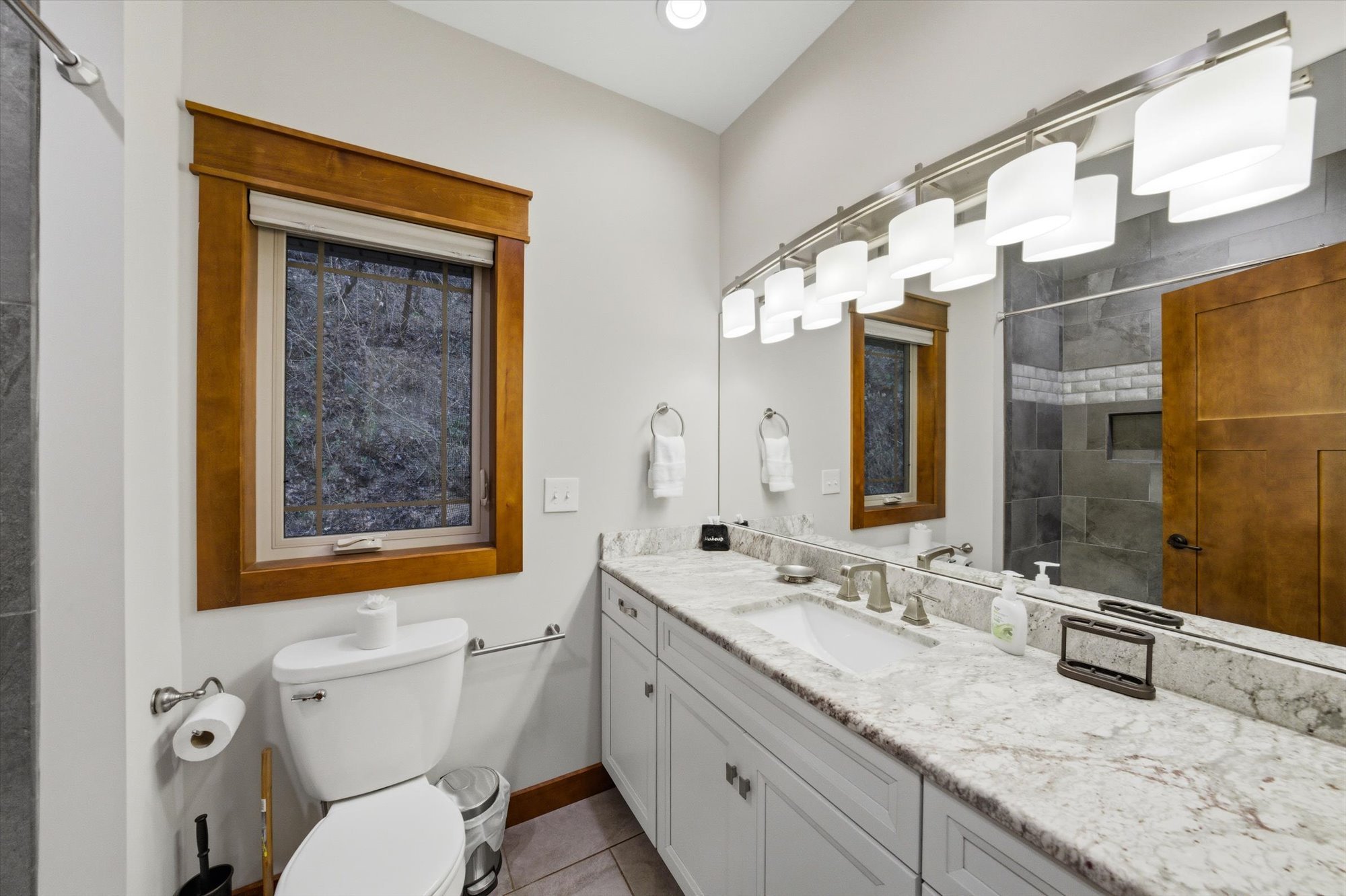 Bathroom with white vanity, granite countertop, large mirror, two windows with wood trim, white toilet, gray tiled shower area, and modern lighting fixture.
