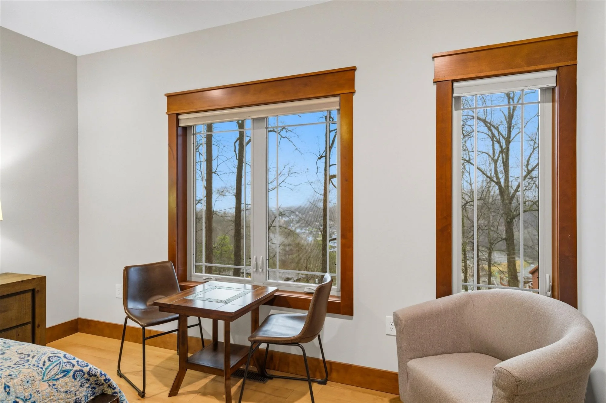 A cozy living room corner featuring two large windows with wooden frames looking out on trees, a small wooden table with two leather chairs, and an upholstered beige armchair.
