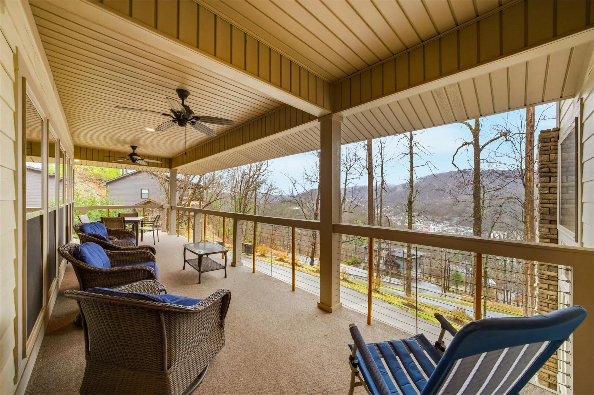 Covered balcony with wicker chairs, blue cushions, a small table, and a view of leafless trees and hills in the distance.