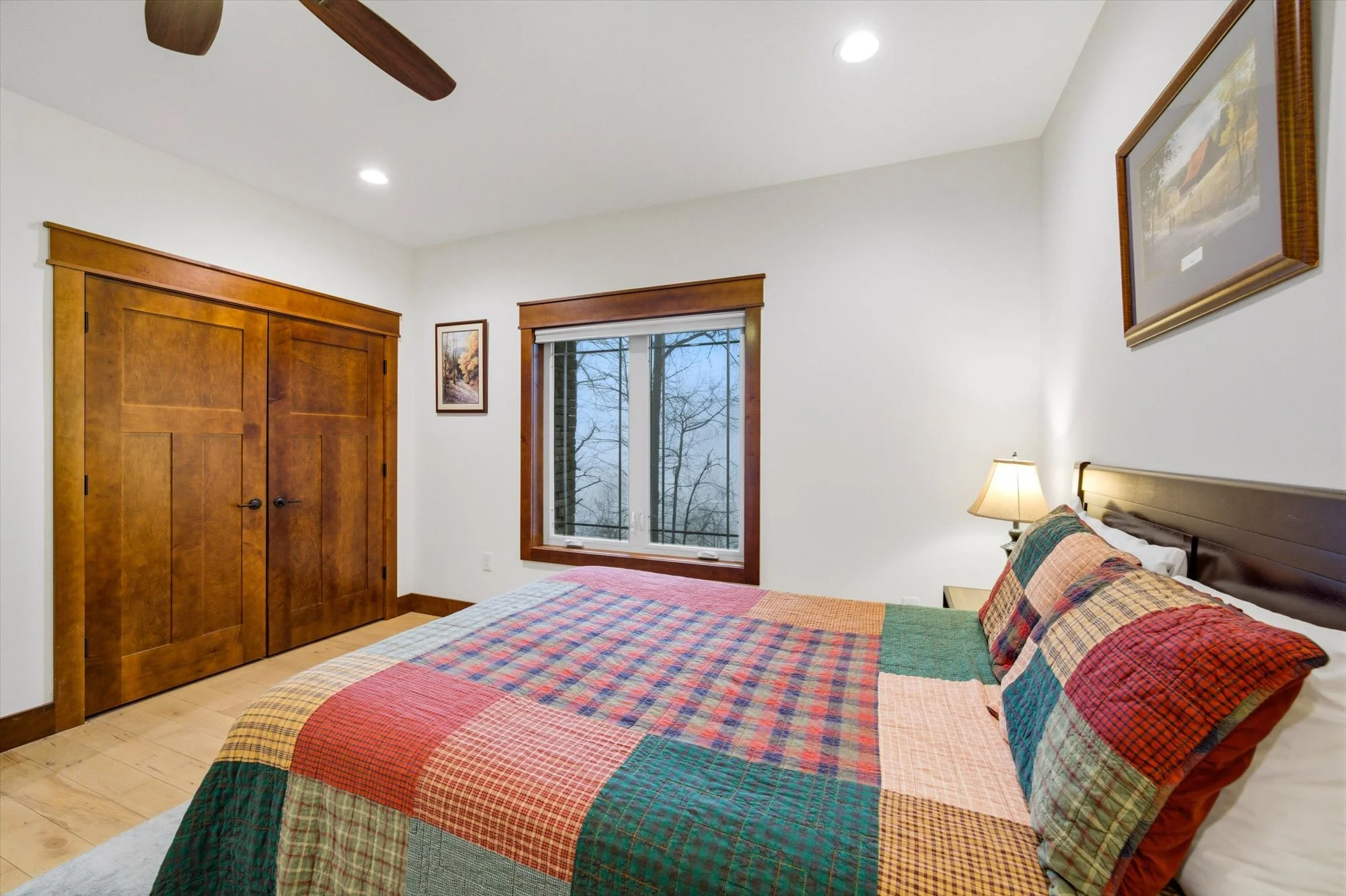 A bedroom with a wooden bed frame, colorful patchwork quilt, a nightstand with a lamp, a window with blinds, and wooden closet doors.