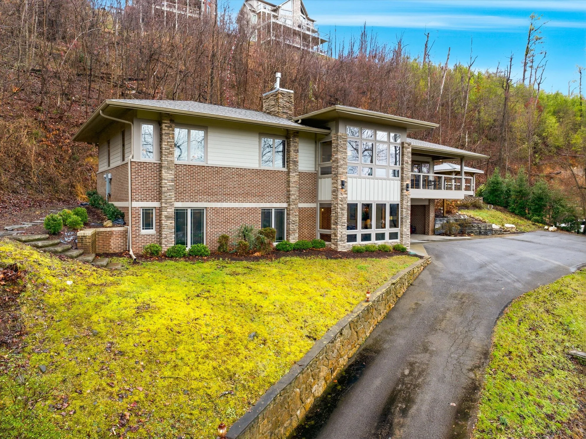 A modern two-story house with brick and white siding exterior, large windows, and a driveway on a slightly sloped, grassy landscape surrounded by trees.