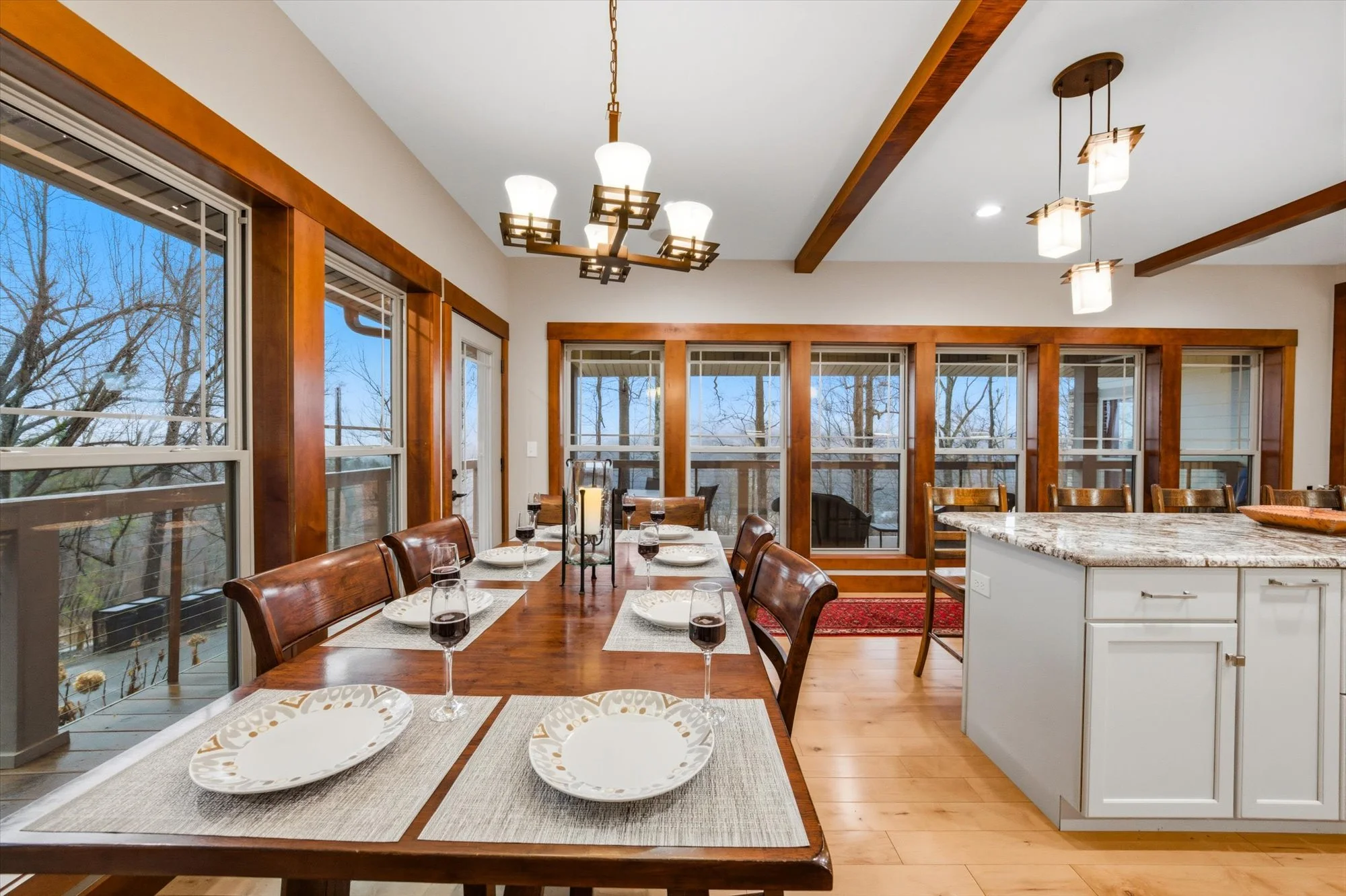 A dining room with a wooden table set with plates and wine glasses, surrounded by wooden chairs, large windows with wooden frames, and a kitchen island with a granite countertop.