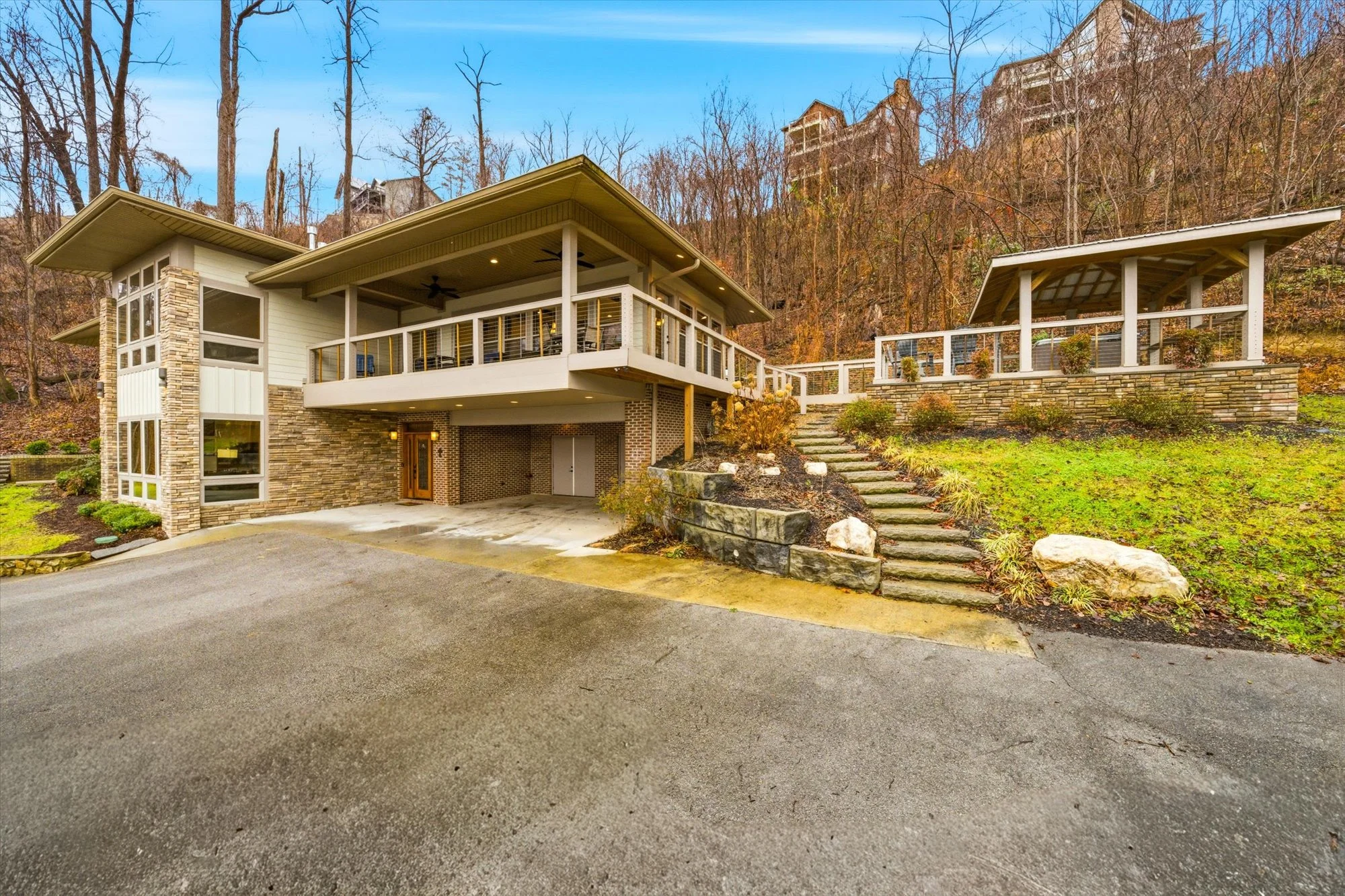 Modern multi-story house with brick and stone exterior, multiple windows, large balcony, and a landscaped front yard with stone steps, surrounded by leafless trees and other homes on a hillside.