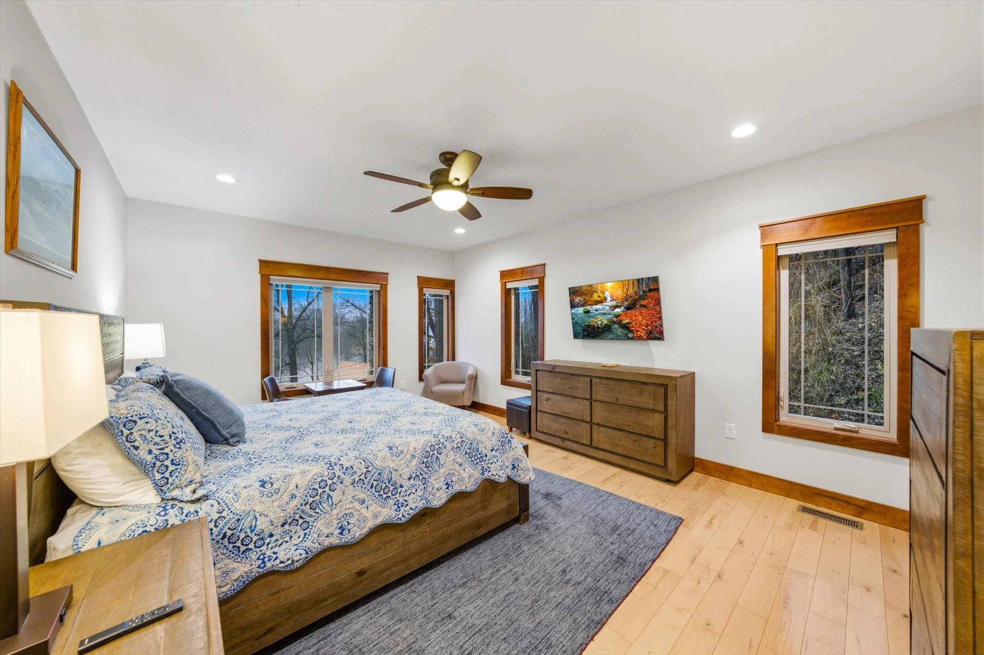 A bedroom with a wooden bed, blue patterned bedding, a window seat, a dresser with a TV, and three windows showing a wooded outdoor scene.
