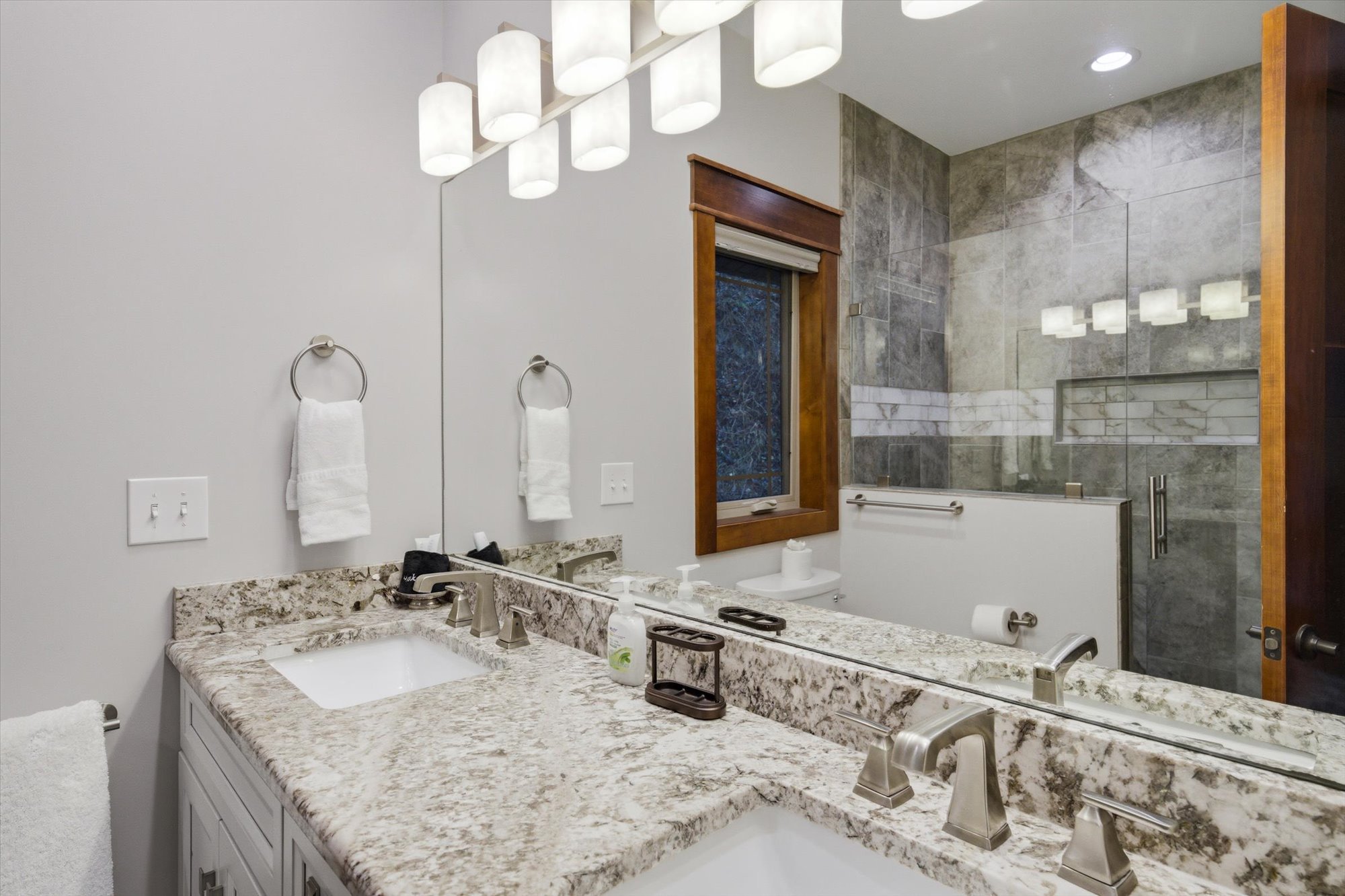Bathroom with granite countertop, double sink, large mirror, towel rings with white towels, soap dispenser, and a window overlooking a tiled shower area with a glass door and grab bar.
