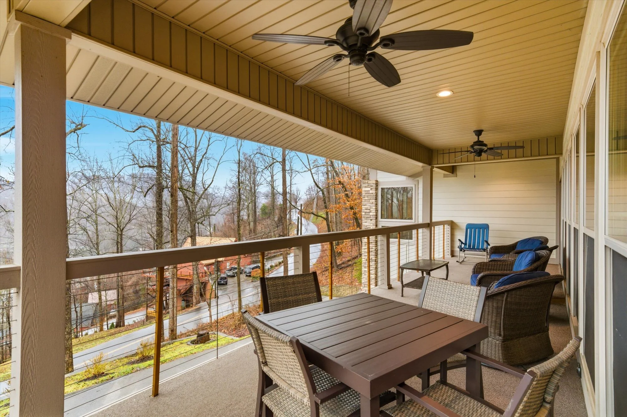 Covered balcony with wooden railing, outdoor furniture including a rectangular dining table, six chairs, and cushioned armchairs, ceiling fans, and a view of a street with trees and houses.