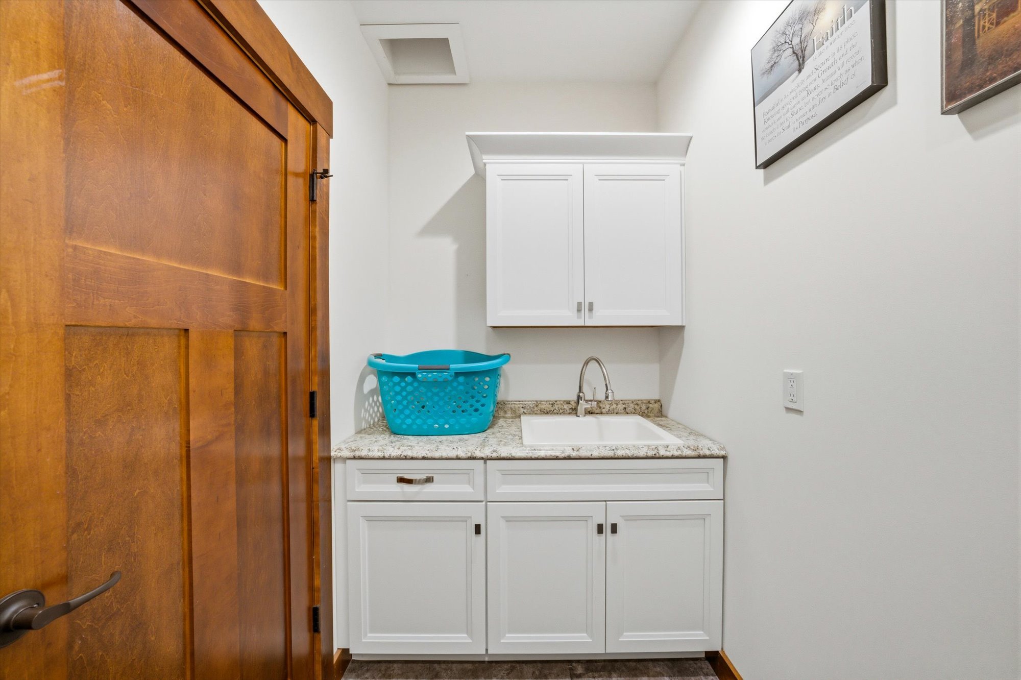 Laundry room with a wooden door, white cabinets above and below a granite countertop, a small sink, and a blue plastic laundry basket on the counter. Two paintings hang on the wall.