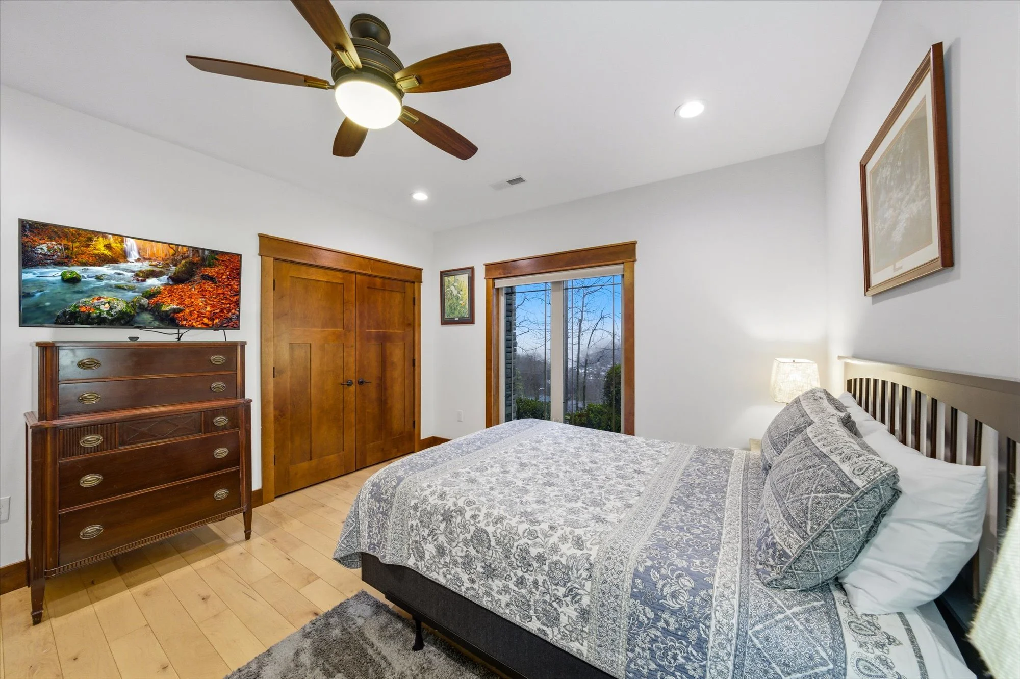 Bedroom with a bed, wooden dresser topped with a television, sliding glass door showing outdoor trees, ceiling fan with wooden blades, and framed art on the walls.