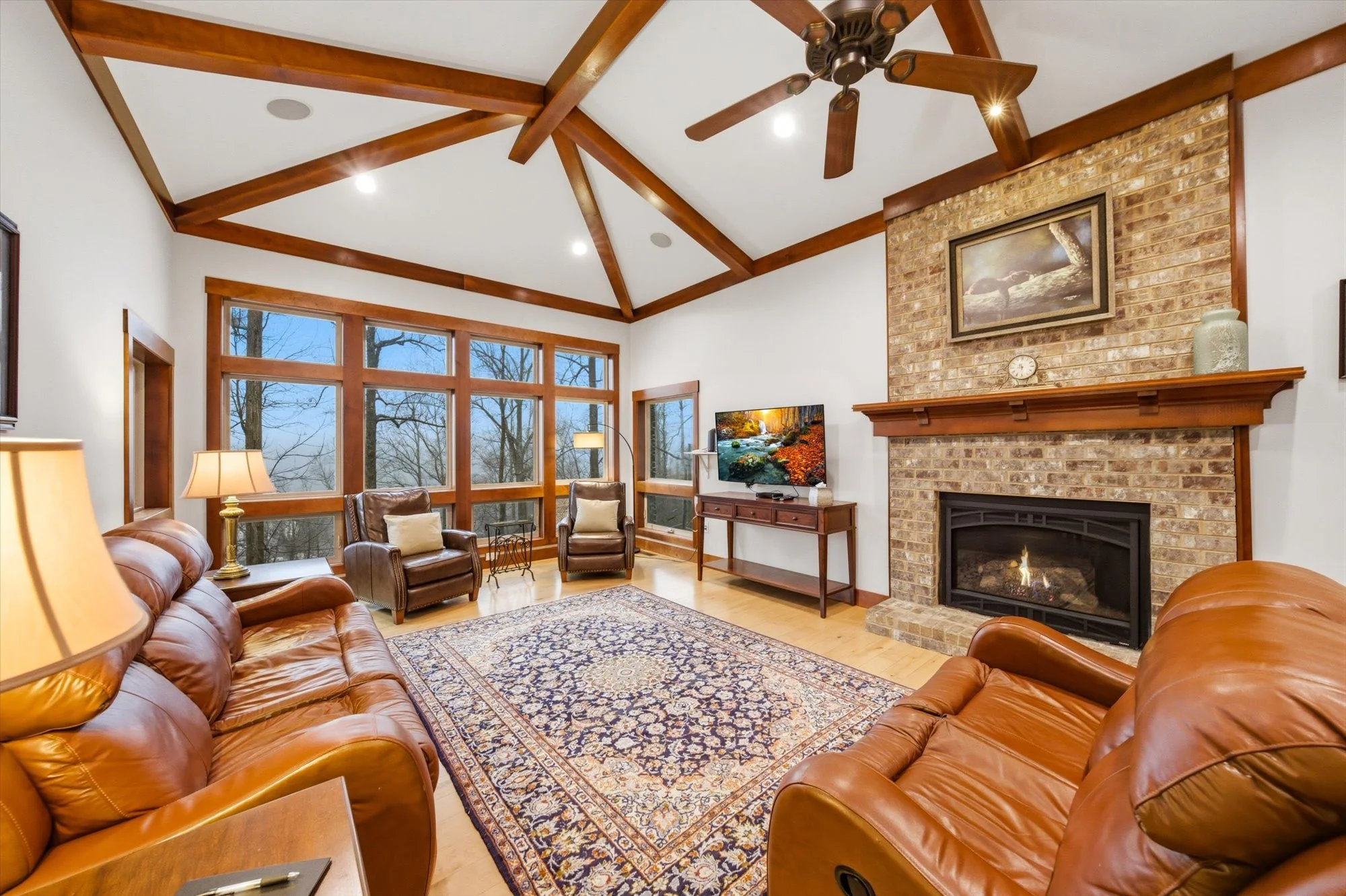 Cozy living room with leather sofas and armchairs, a large patterned area rug, brick fireplace with a wooden mantle, a framed picture above, a flat-screen TV, large windows with a view of leafless trees, ceiling fan, and recessed lighting.