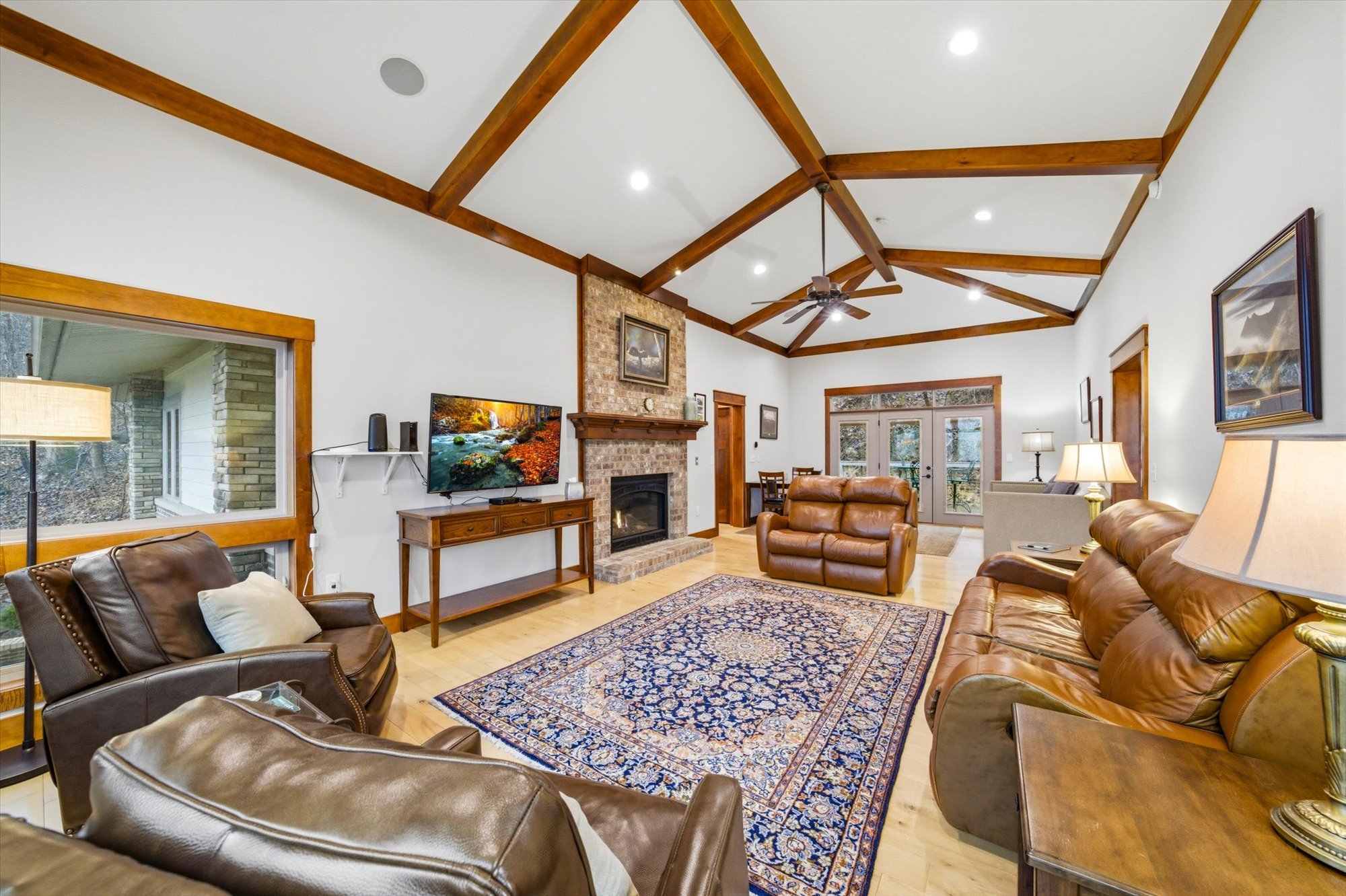 Spacious living room with wood beam ceiling, brick fireplace, leather sofas, a rug, large windows, and a TV.