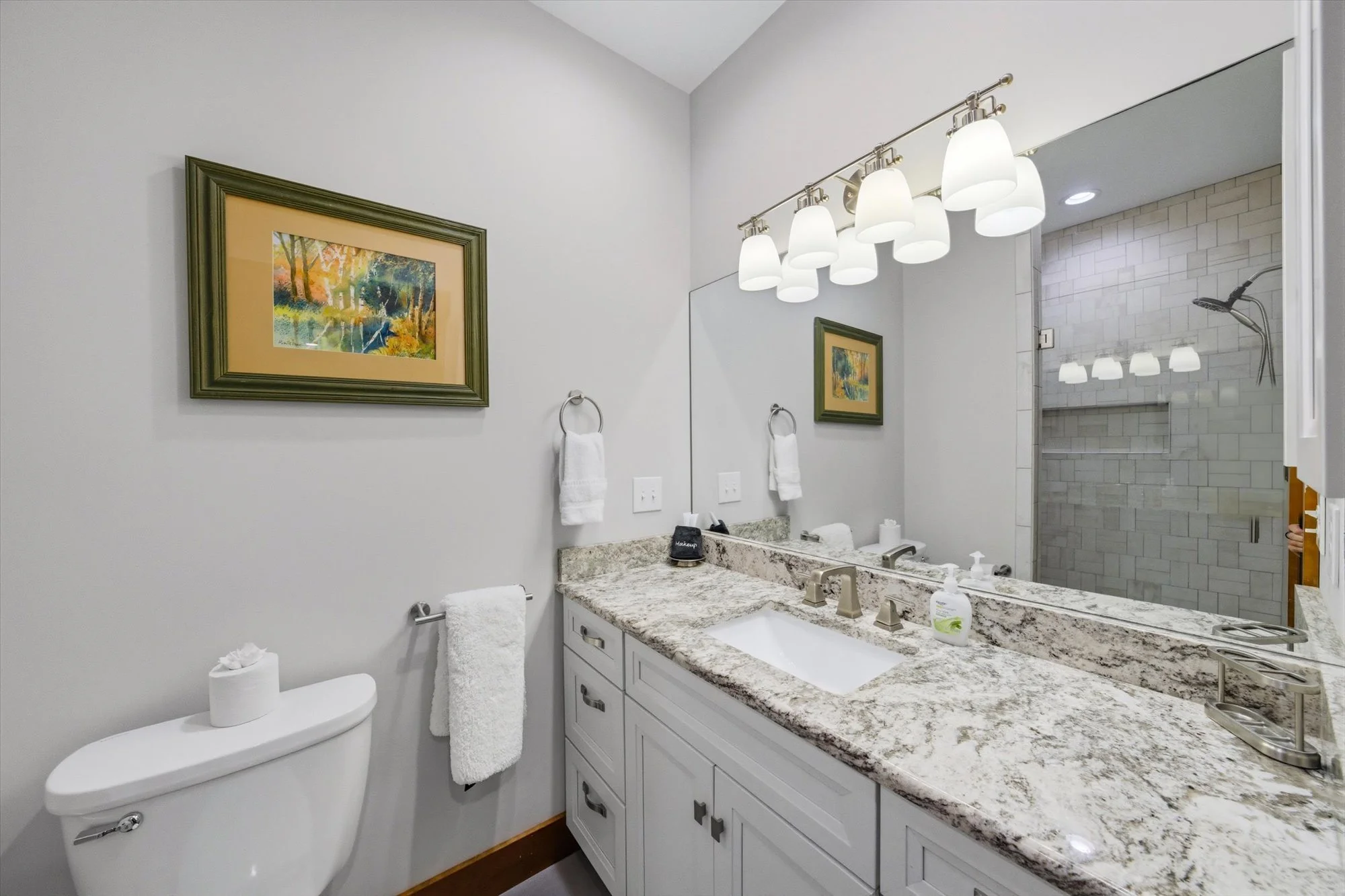 Bathroom with a granite countertop vanity, large mirror, white cabinets, a framed landscape painting, a white toilet with a decorative tissue holder, two towels hanging on the wall, a soap dispenser, and a walk-in shower with a glass door and gray ti