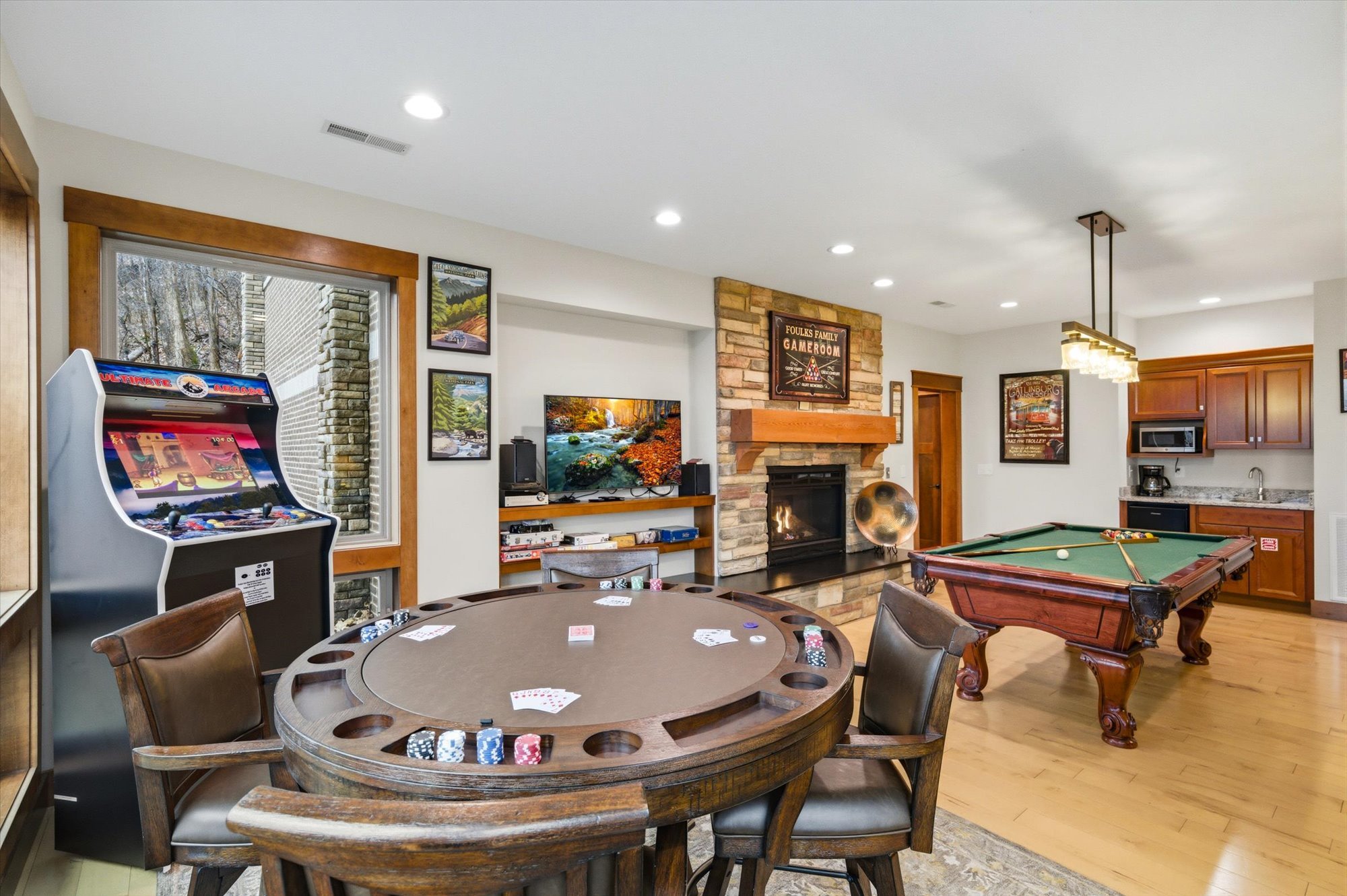A game room featuring a poker table with chips and cards, a billiards table, a fireplace with a brick chimney, a flat-screen TV, an arcade machine, wood cabinets, and wall art.