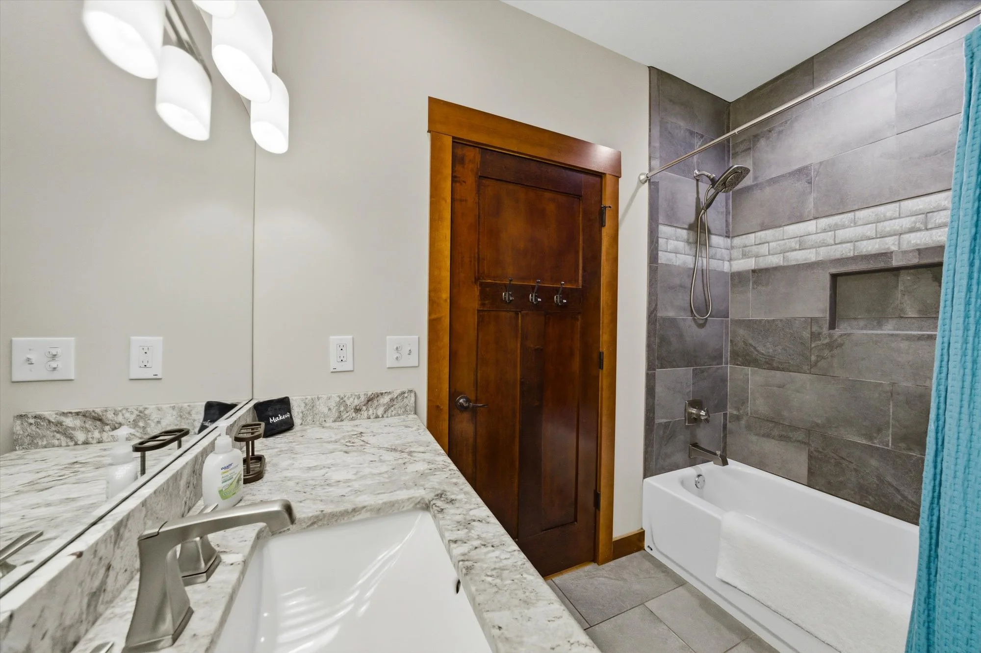 Bathroom with a granite countertop sink, a mirror, wall lights, a wooden door, a bathtub with a tiled wall, and a blue towel hanging on a shower rod.