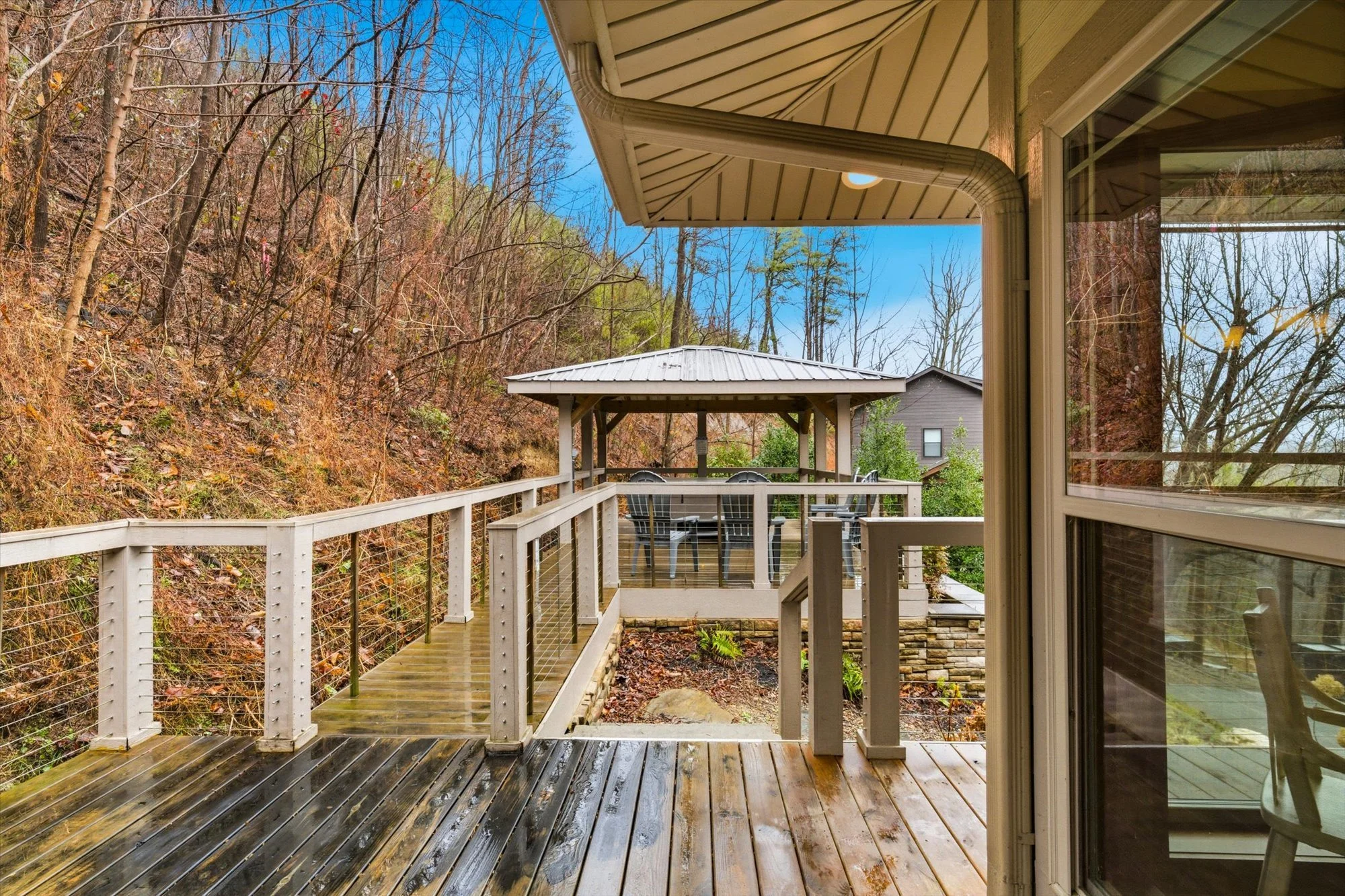 View from a house's sliding glass door showing a wooden deck with a gazebo and outdoor dining table, overlooking a wooded hillside with leafless trees under a bright sky.