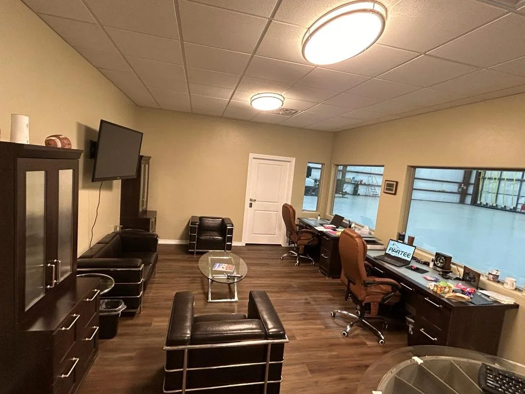 Office room with three desks, one with a computer monitor, two with laptops, and various office supplies. There are two brown chairs, a black leather sofa, a glass coffee table, and a wall-mounted flat-screen TV. The room has wood flooring and large 