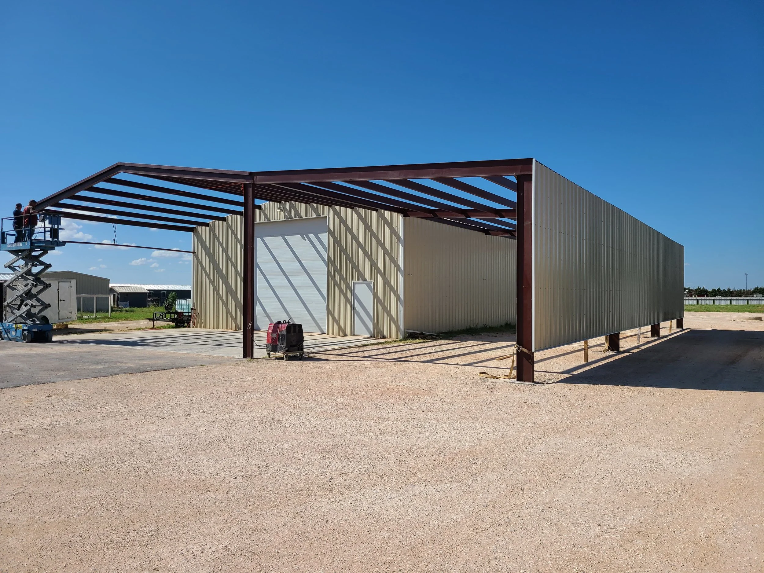 Under construction metal building with roof framework, a scissor lift on the left with workers, a portable heater, and a dirt and paved area in the foreground.