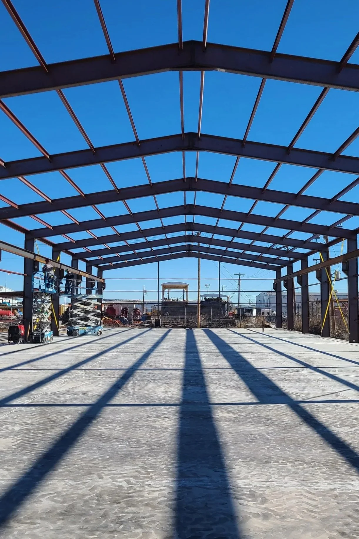 Steel framework structure of a building under construction with shadows on concrete floor, and construction equipment on the sides, set against a clear blue sky.