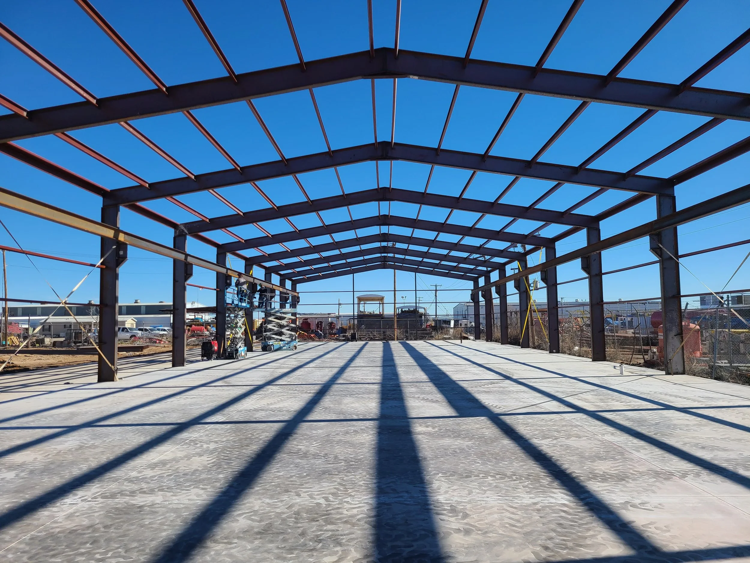 Construction site with a steel frame structure and concrete floor, with shadows cast by the beams under a clear blue sky.