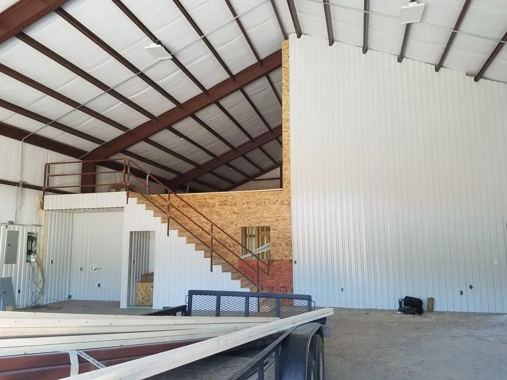 Interior of a building under construction featuring a metal roof, exposed wooden framing, a staircase with metal railing, and partially finished wall panels.