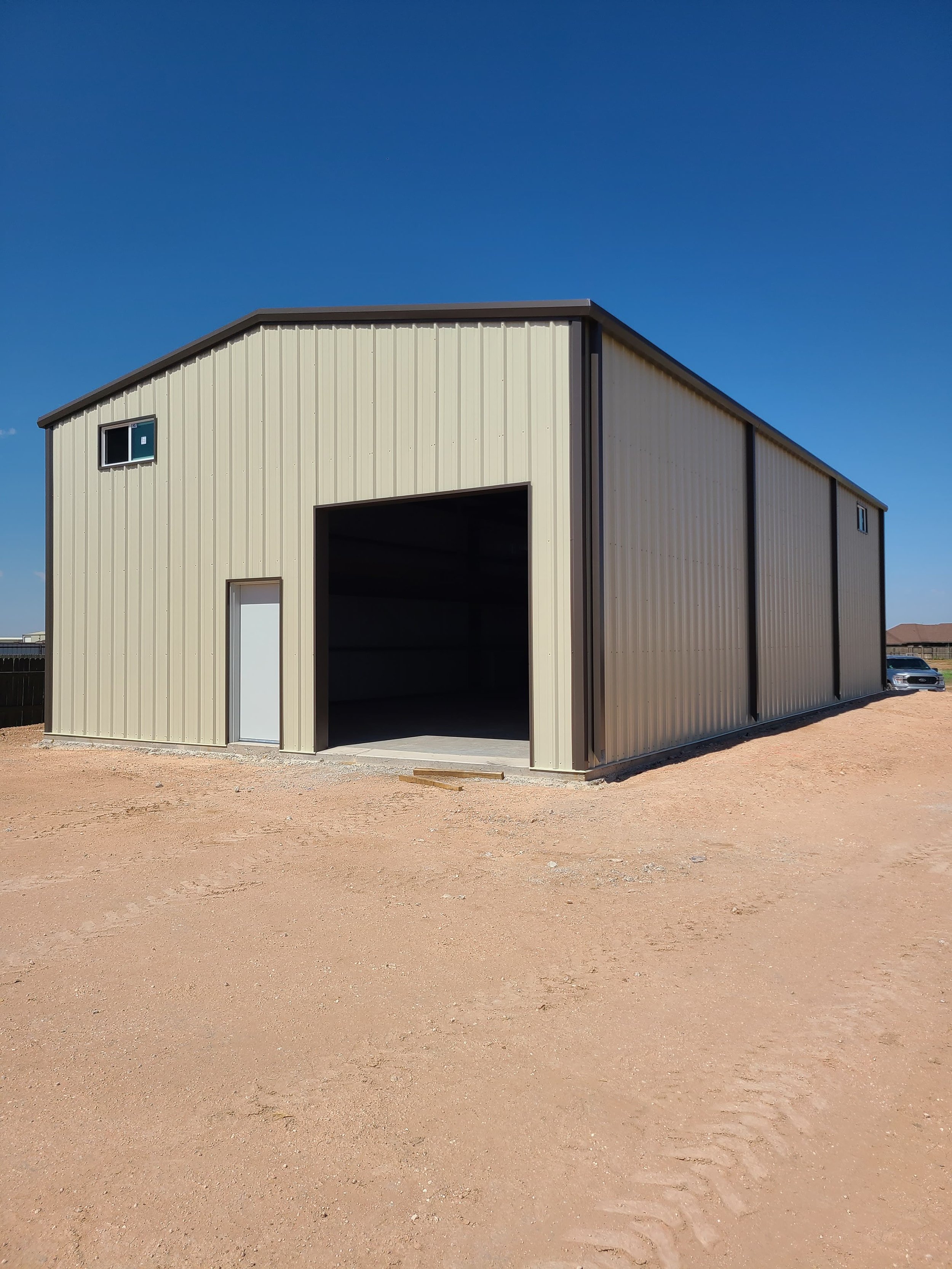 A beige metal storage building with a large open door and a smaller closed door, situated on a dirt lot under a clear blue sky.