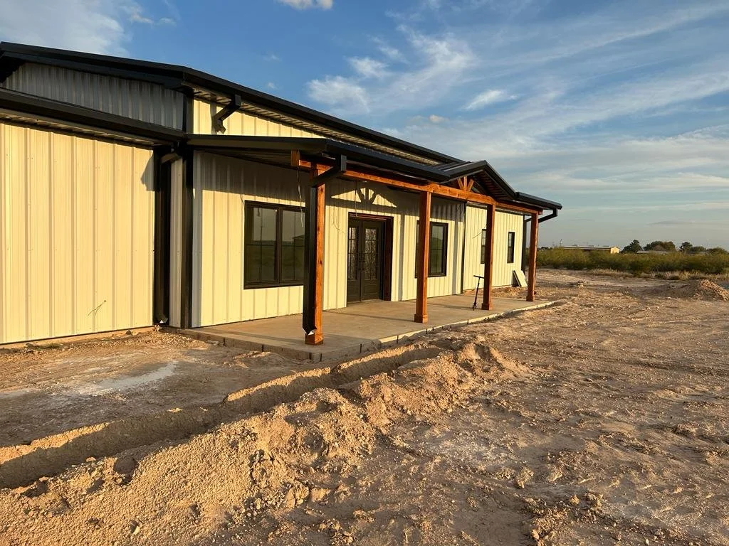 New metal building with wooden porch in rural area, partly built with dirt ground in foreground, under a partly cloudy sky.