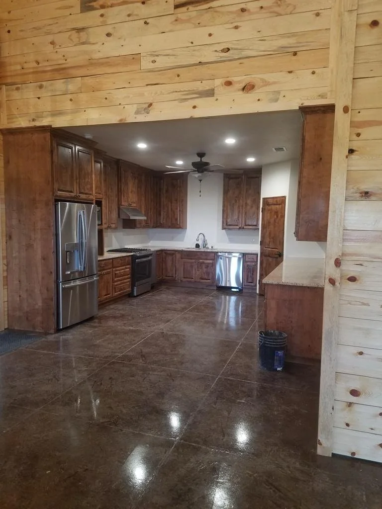 Empty modern kitchen with wooden cabinets, stainless steel appliances, and a polished concrete floor, with wood-paneled walls and ceiling fan.