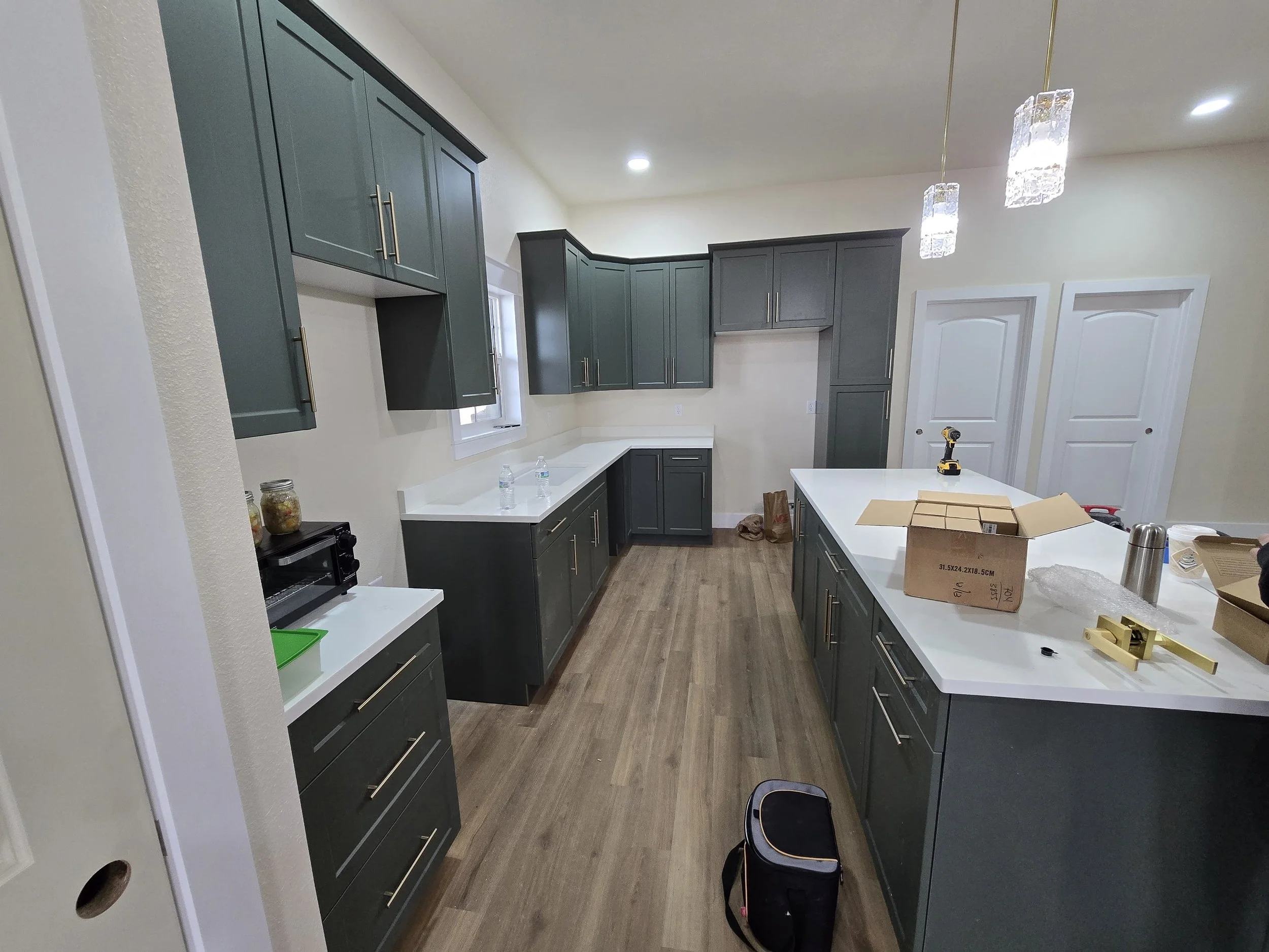 Modern kitchen with dark gray cabinets, island with various tools and boxes, hardwood floor, pendant lights, and double white doors.