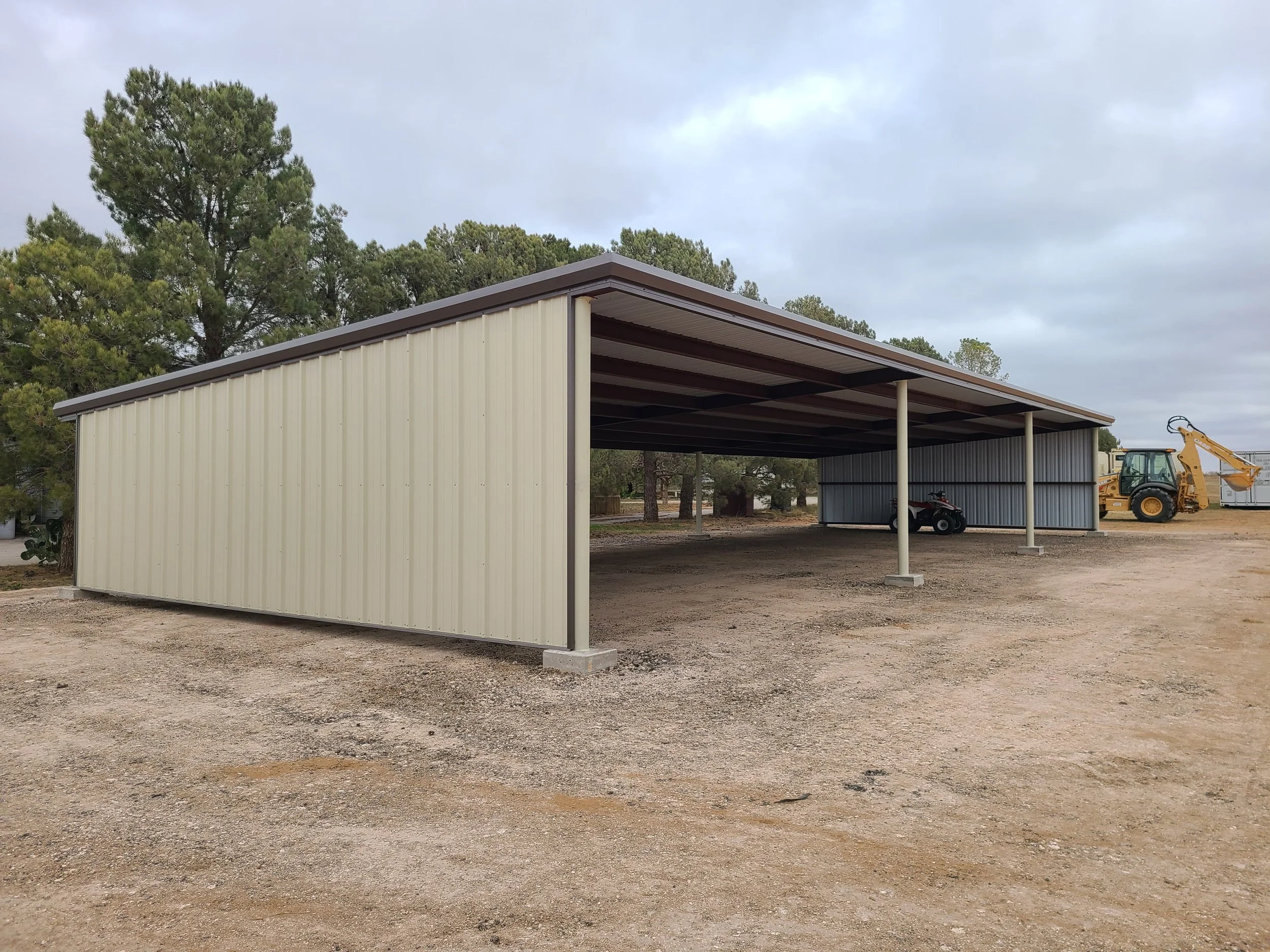 A beige metal carport with a brown roof, supported by metal poles, situated on a gravel lot with trees in the background. Under the carport are a motorcycle and a small utility vehicle. To the right, there is a yellow backhoe loader and a small shed.