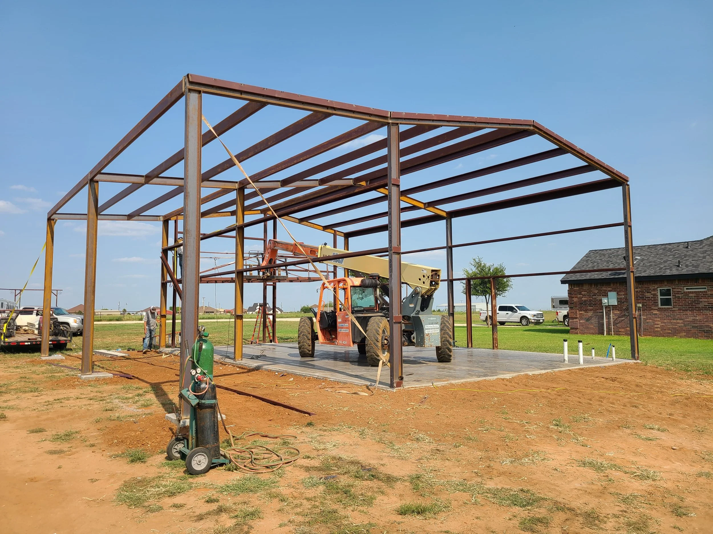 Under construction steel frame building with a forklift on a concrete slab, construction workers, and vehicles in the background.