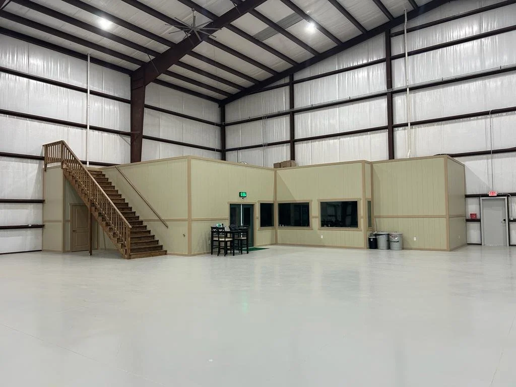 Large empty warehouse with a small office area built inside, featuring a staircase, windows, and trash cans.