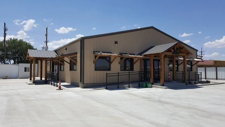 New beige building with black trim, wooden porch with awning, parking lot in foreground, blue sky with few clouds.
