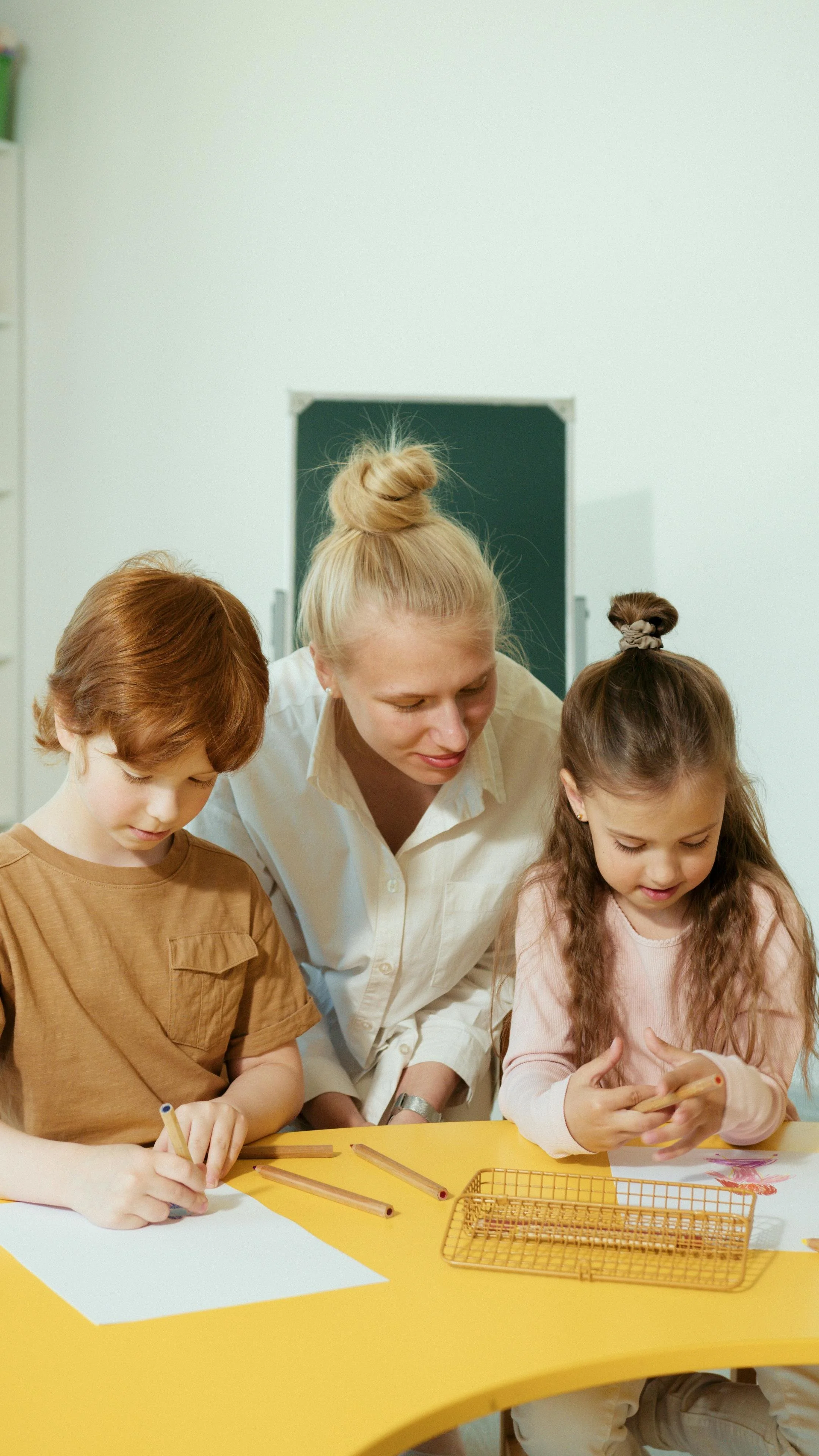 Woman with a hair bun helps two school children with a project.