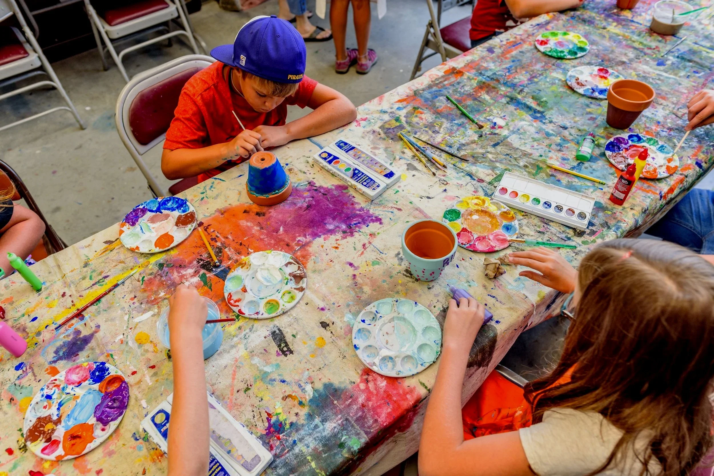 Kids painting on a long table with lots of colors everywhere.