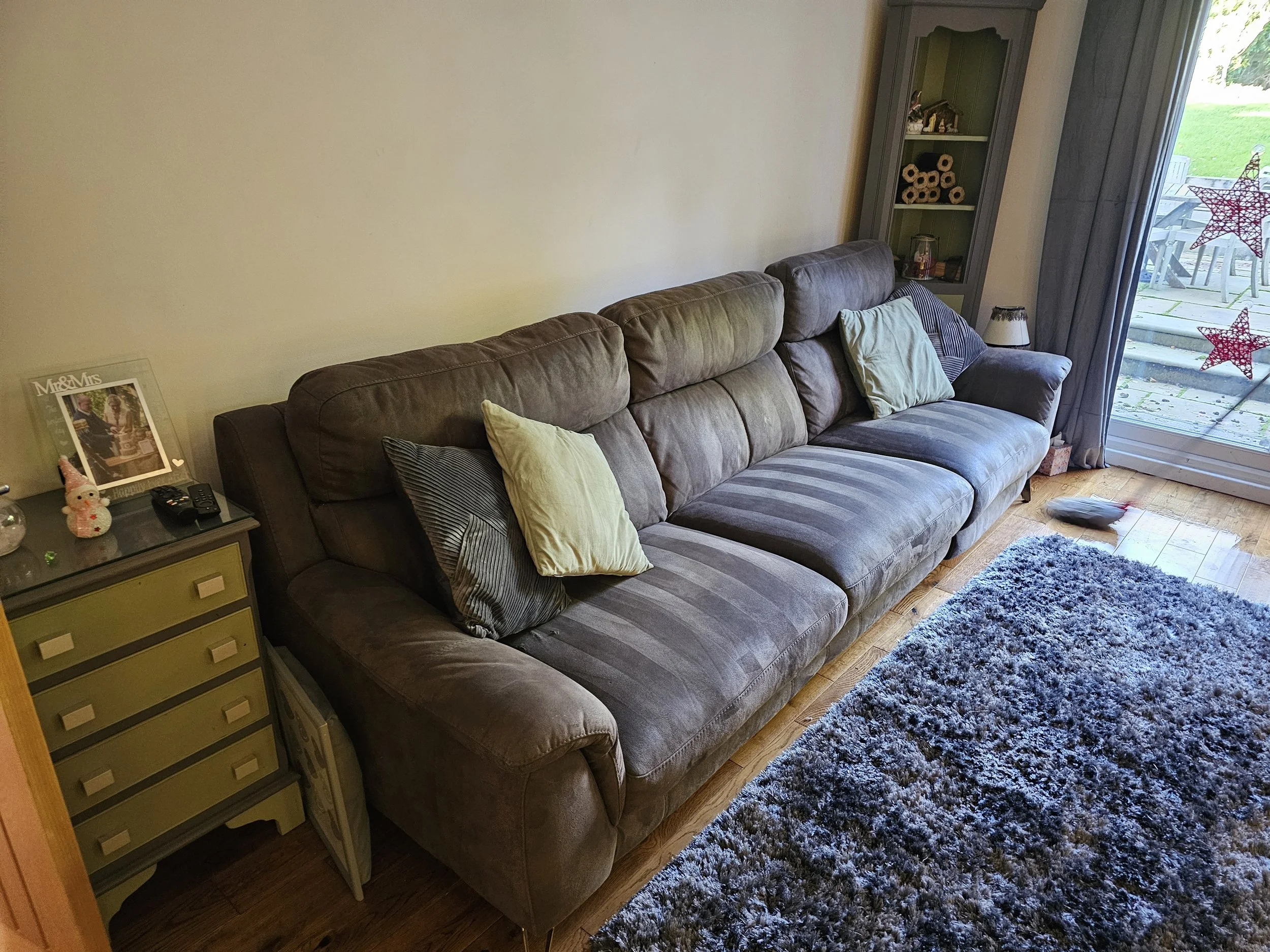 Living room with a large gray sofa, a dark green cabinet, and a sliding glass door with holiday star decorations. There is a gray shaggy rug on a wooden floor and a small side table with a framed photo, a snowman figurine, and a remote control.