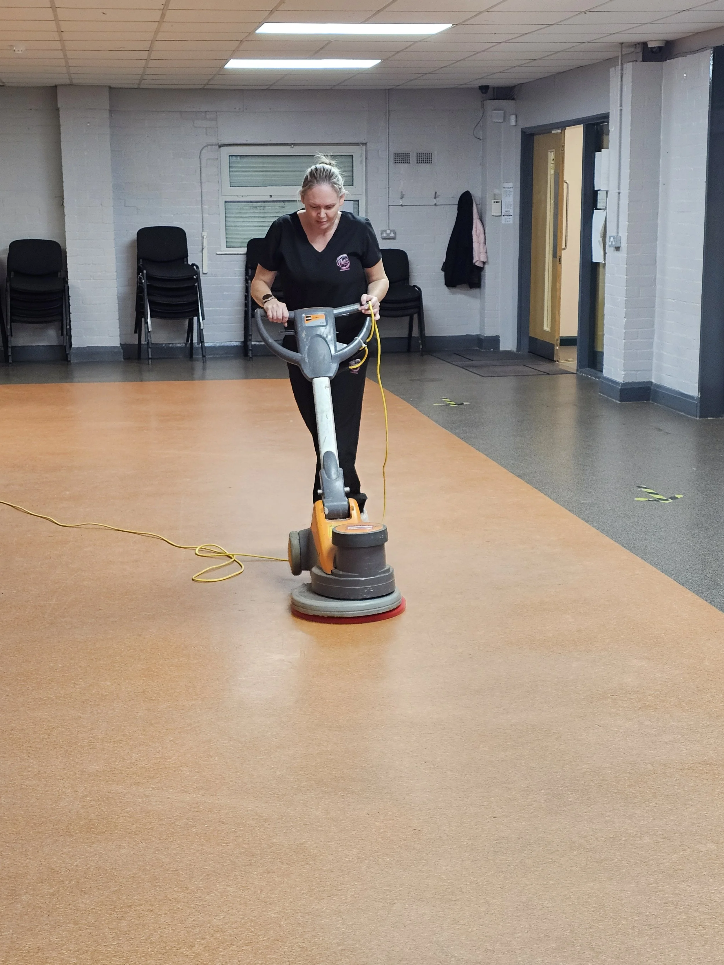 Woman using a floor buffer machine on a tan carpet in a room with white brick walls and black chairs