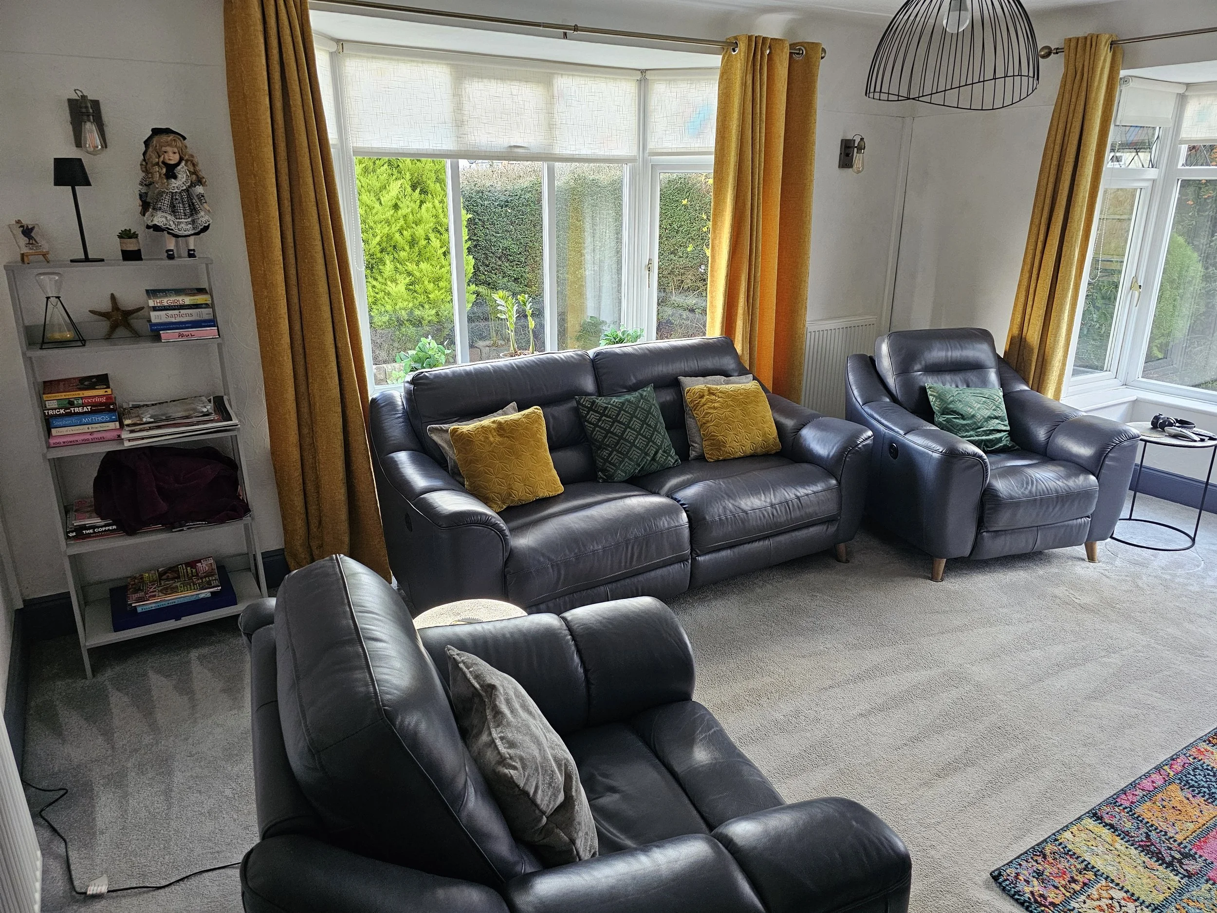 Living room with three black leather armchairs and a black leather sofa with yellow and green cushions, large windows with yellow curtains, a white bookshelf with books, a doll, and decorative items, a colorful rug, and a small side table with remote controls.