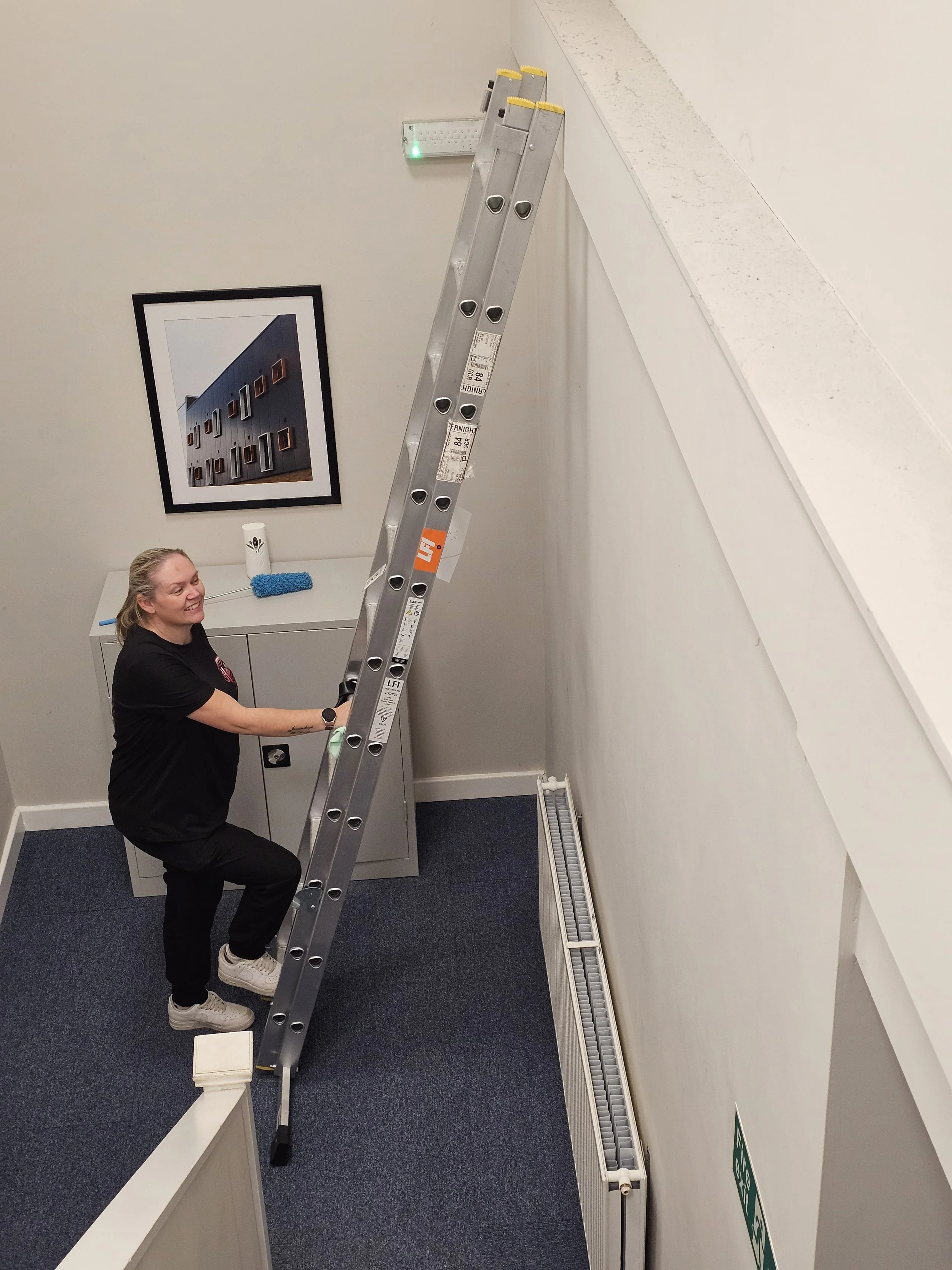 A woman moving a tall ladder in a small office room with artwork on the wall and a locker.