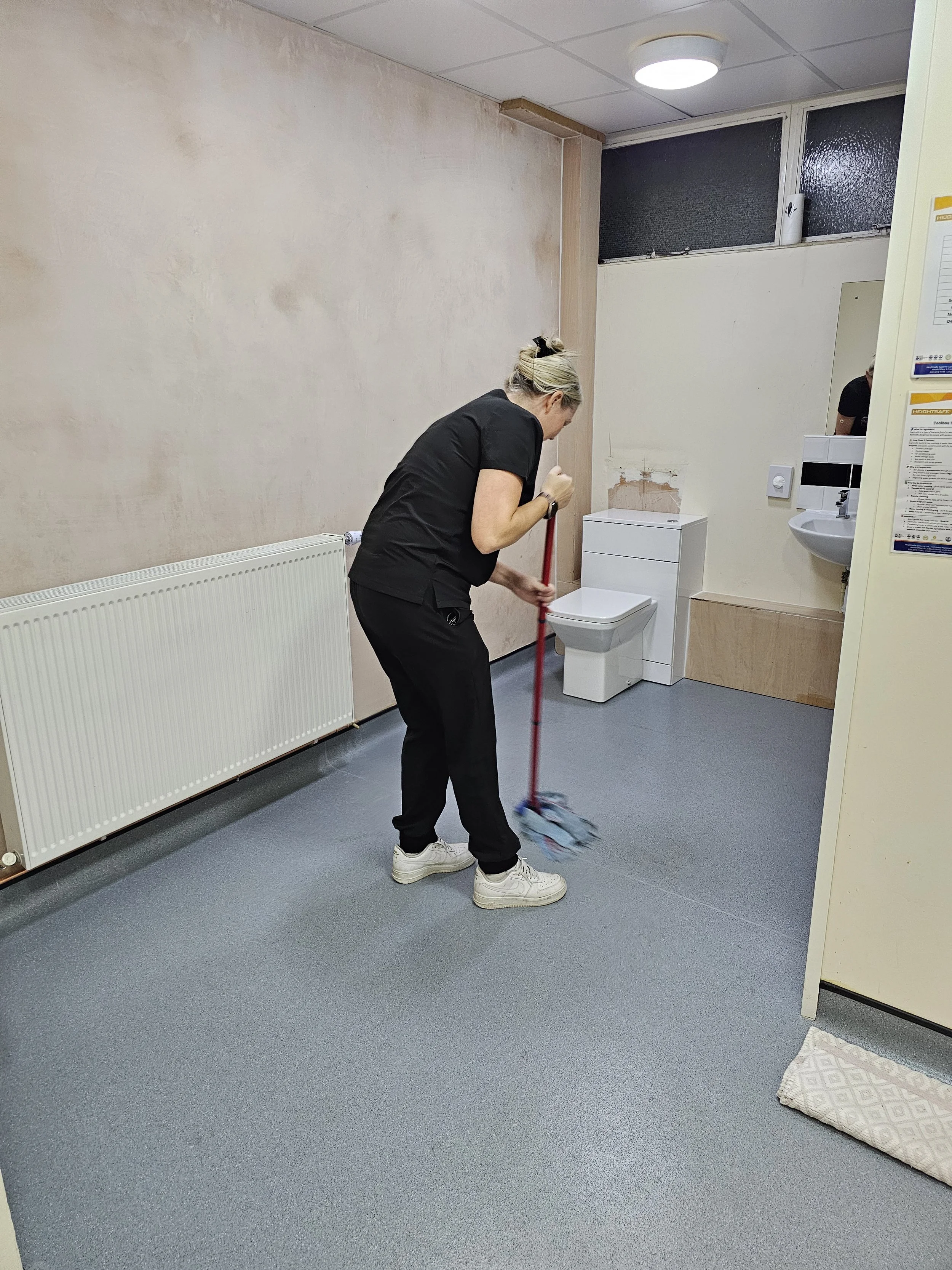 A woman in black scrubs sweeping the floor of a bathroom or laundry room with a mop.